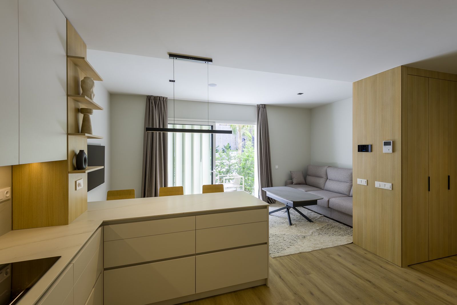 Wide-angle view of a modern apartment interior featuring a white kitchen peninsula with drawers, a light-wood panel wall with built-in storage, a grey fabric sofa over a textured rug, and glass patio doors framed by taupe curtains leading to an outdoor garden.