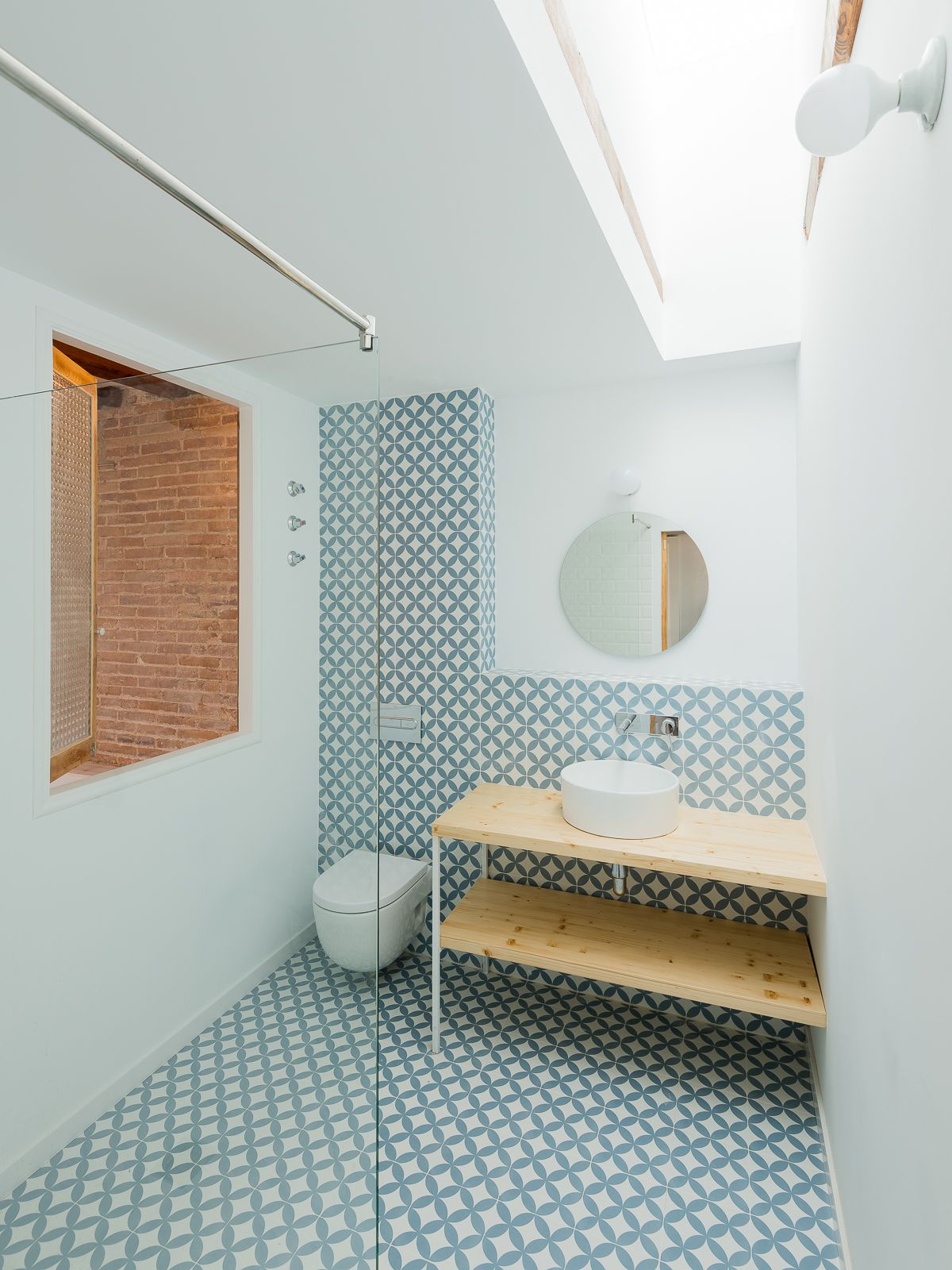 Baño con diseño de baldosas geométricas azules y blancas, espejo redondo, tragaluz y un tocador de madera con lavabo de recipiente.