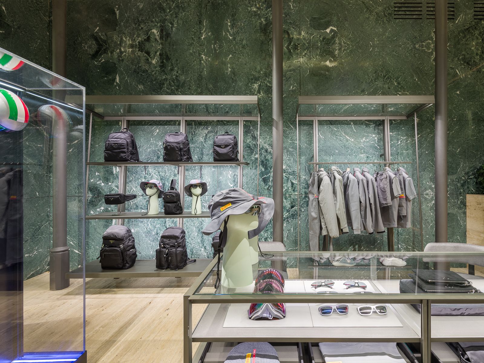 Boutique de lujo con paredes de mármol verde, mostrando zapatillas de diseño, bolsos negros y accesorios en elegantes estanterías metálicas. Una mesa de vidrio exhibe gafas y sombreros, mientras que un maniquí en un atuendo de rendimiento está encerrado en una vitrina de vidrio.