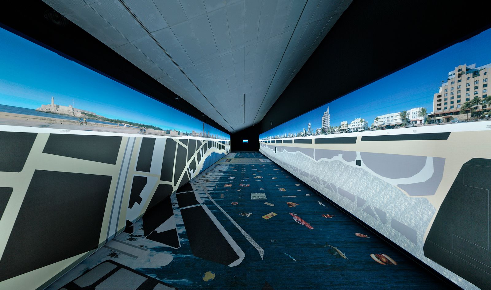 Una sala de exposición larga y estrecha con imágenes panorámicas de un paisaje urbano y playa mostradas en las paredes, con planos arquitectónicos detallados y mapas en el suelo.