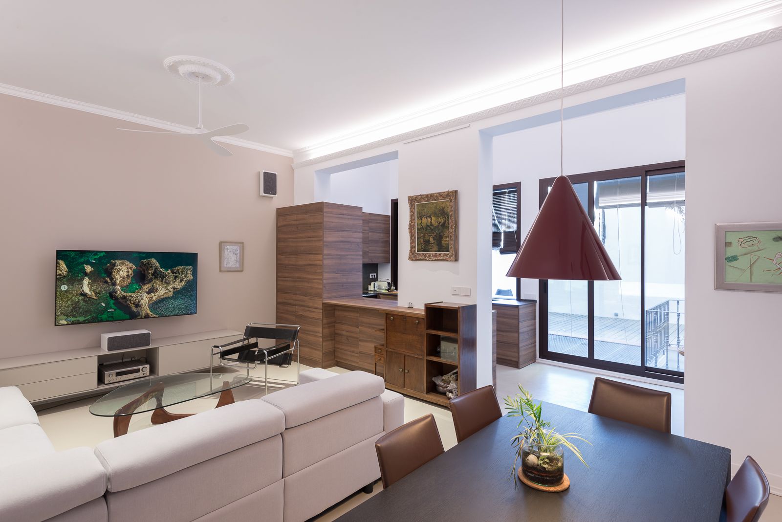 Modern living room with a large TV, white sectional sofa, glass coffee table, and dining area. Patio access through sliding glass doors.