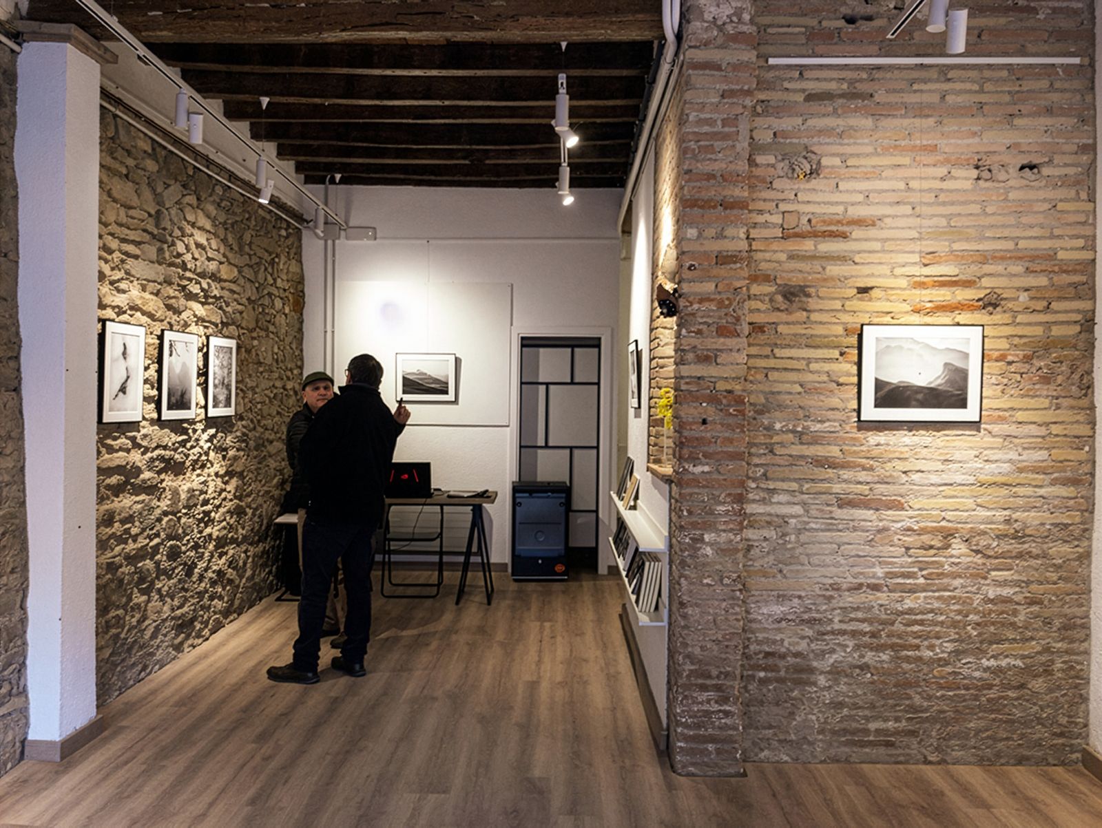Dos personas conversando en una exposición de fotografía con fotos de paisajes en blanco y negro en paredes de piedra y ladrillo.