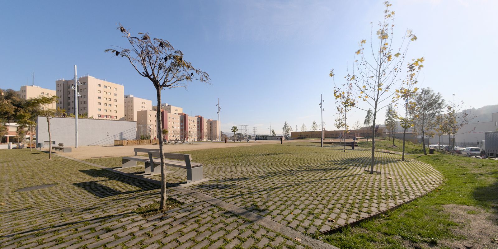 A low-angle landscape photograph of an urban public space at Foneria Park. The foreground features a large expanse of lattice concrete grass pavers, minimalist wooden and metal benches, and slender, young trees. In the background, multi-story residential buildings line the perimeter under a bright, clear blue sky.