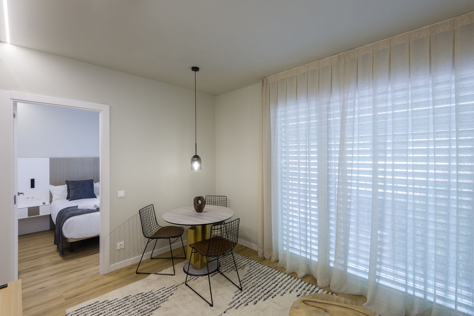 Modern apartment dining nook featuring a small round wooden table with black wire mesh chairs, set over a patterned cream area rug beside floor-to-ceiling sheer white curtains, with a view into an adjacent minimalist bedroom.