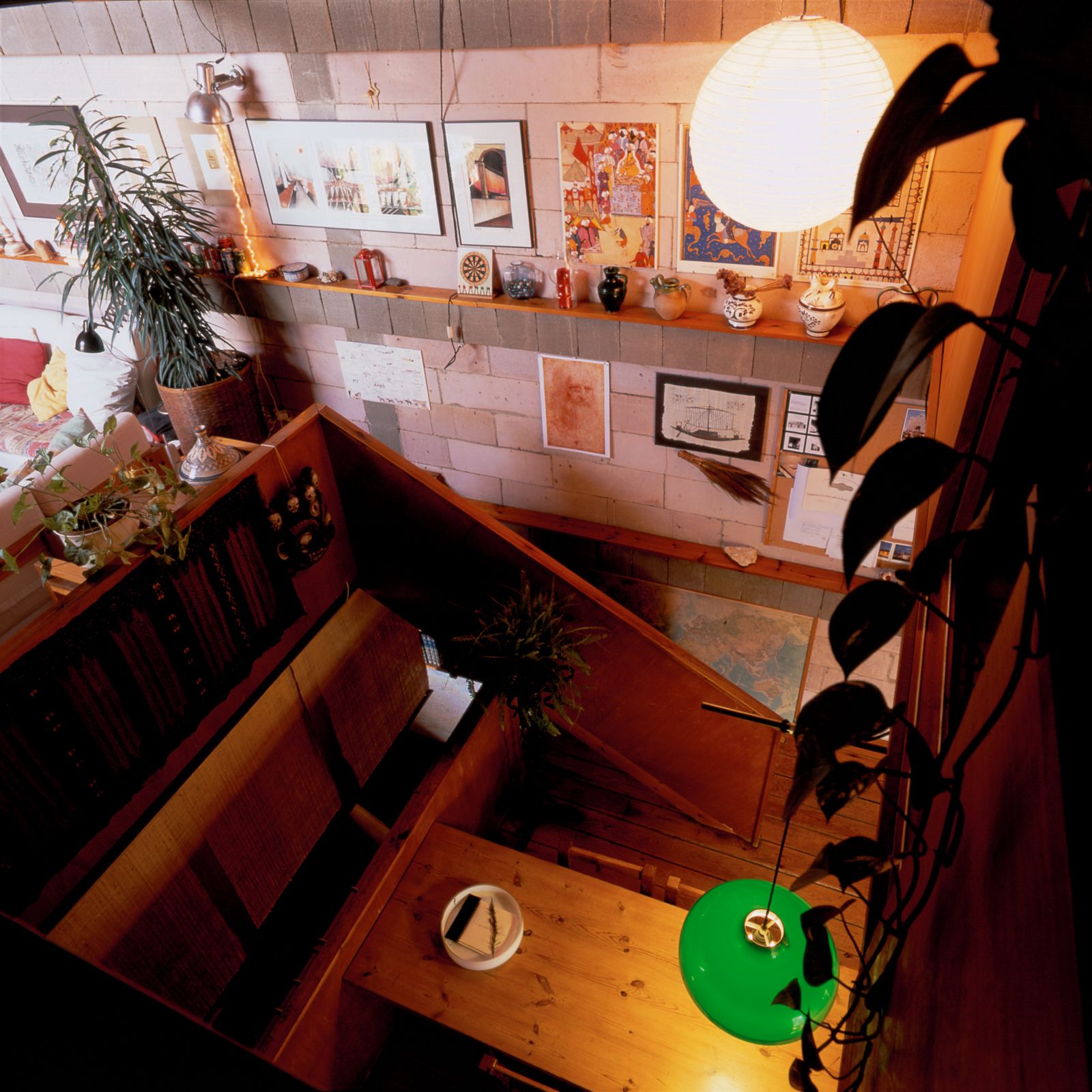 Vista cenital de una acogedora zona de estar con una escalera de madera, una lámpara colgante verde y una pared adornada con arte enmarcado y objetos decorativos. Debajo se puede ver una gran mesa de madera.