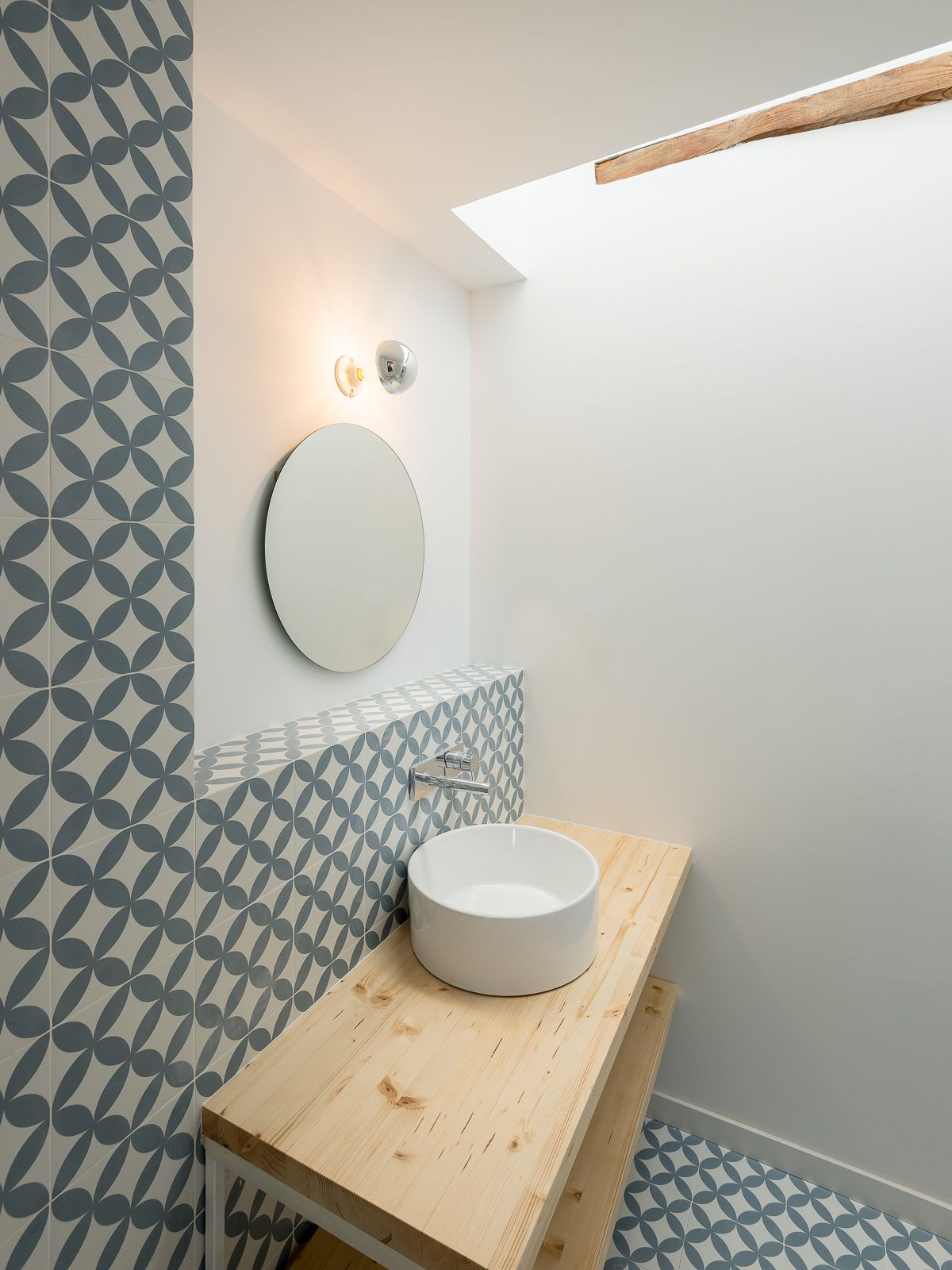 Baño con espejo redondo, lavabo de recipiente, tocador de madera y baldosas geométricas azules y blancas, iluminado por luz natural de un tragaluz.