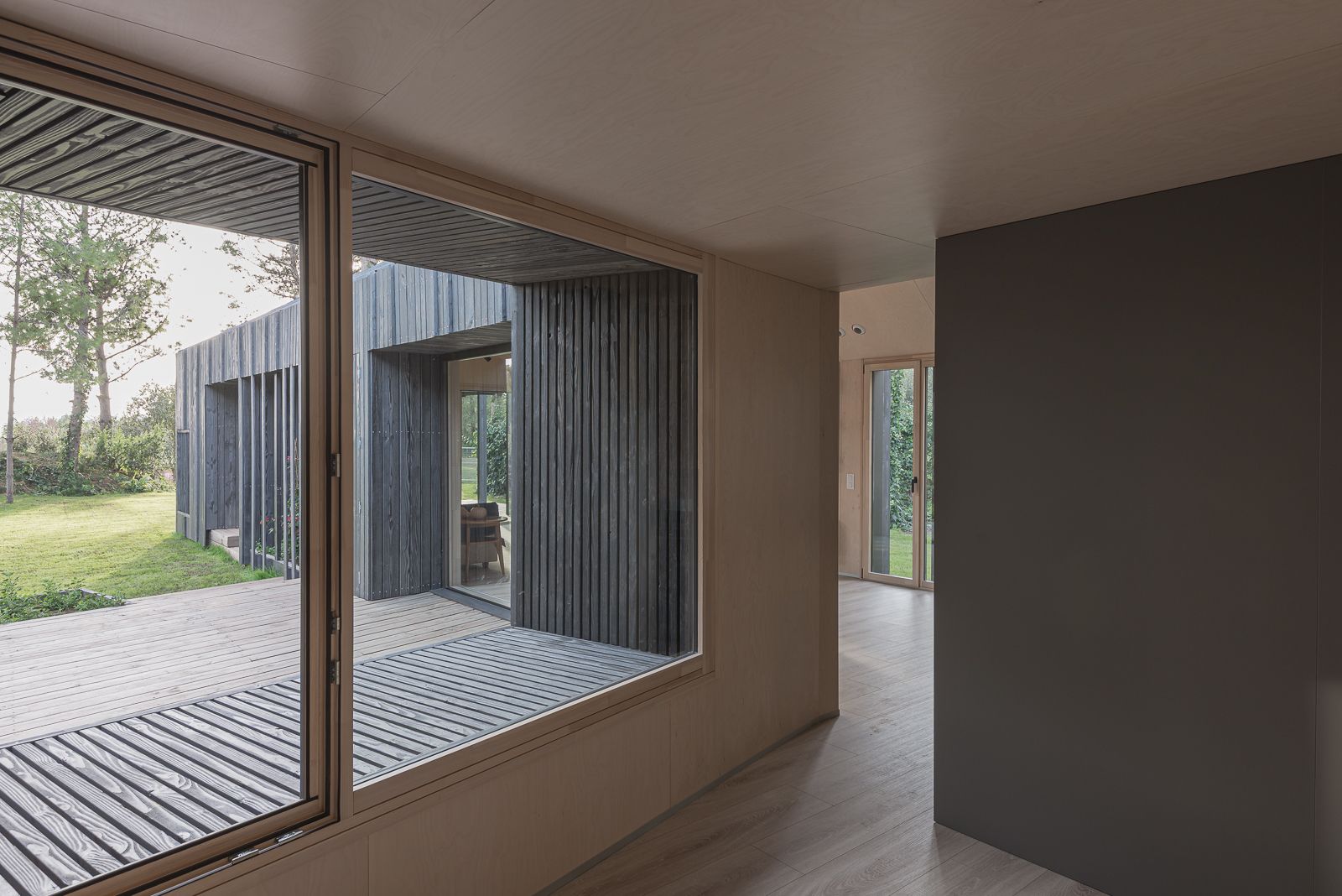 Vista interior de un pasillo con grandes ventanas con vistas a una terraza de madera y área de jardín, mostrando el diseño moderno y la conexión entre los espacios interiores y exteriores.