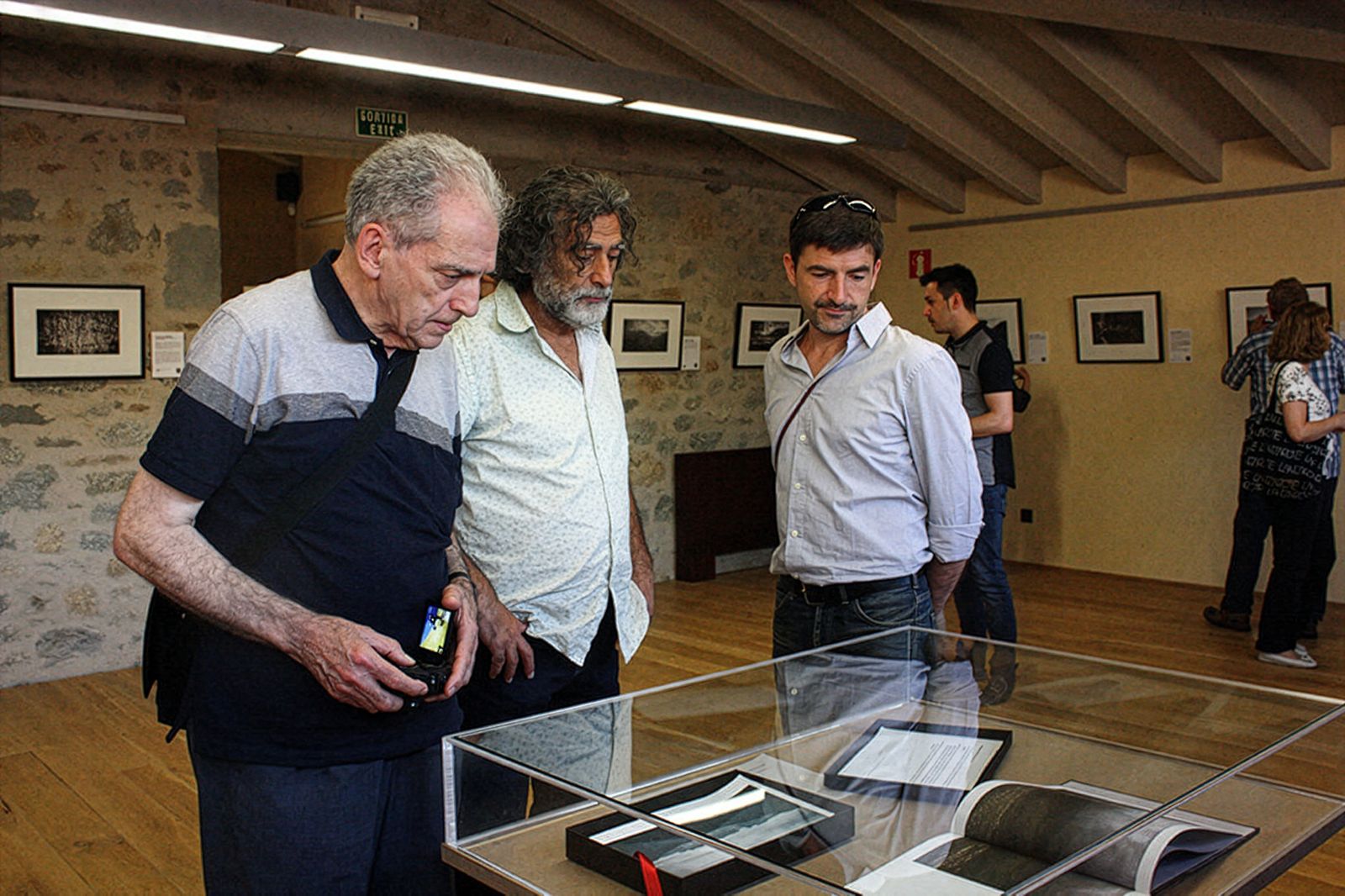 Tres hombres mirando una exhibición en una vitrina en una sala con fotos enmarcadas en paredes de piedra.