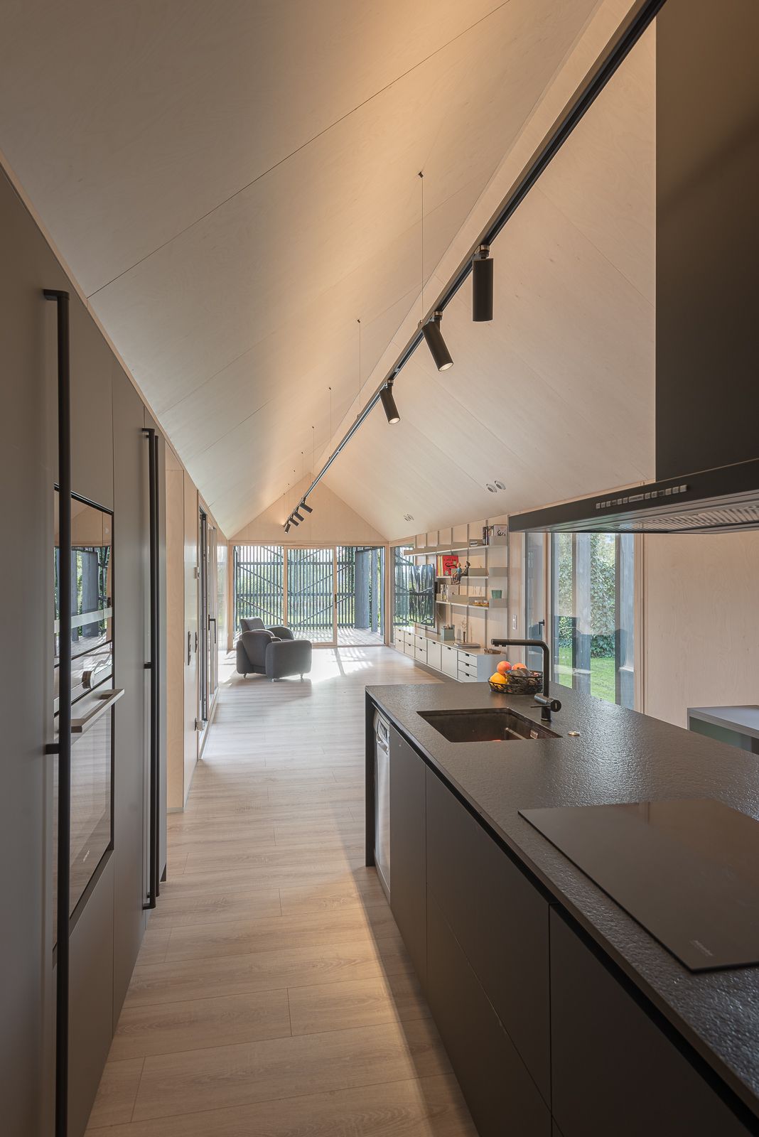 Vista de una cocina moderna con una elegante isla negra con fregadero integrado y encimera de cocción, suelo de madera clara y un techo alto inclinado con iluminación de riel. Grandes ventanas y puertas de vidrio proporcionan abundante luz natural y vistas de la vegetación exterior.