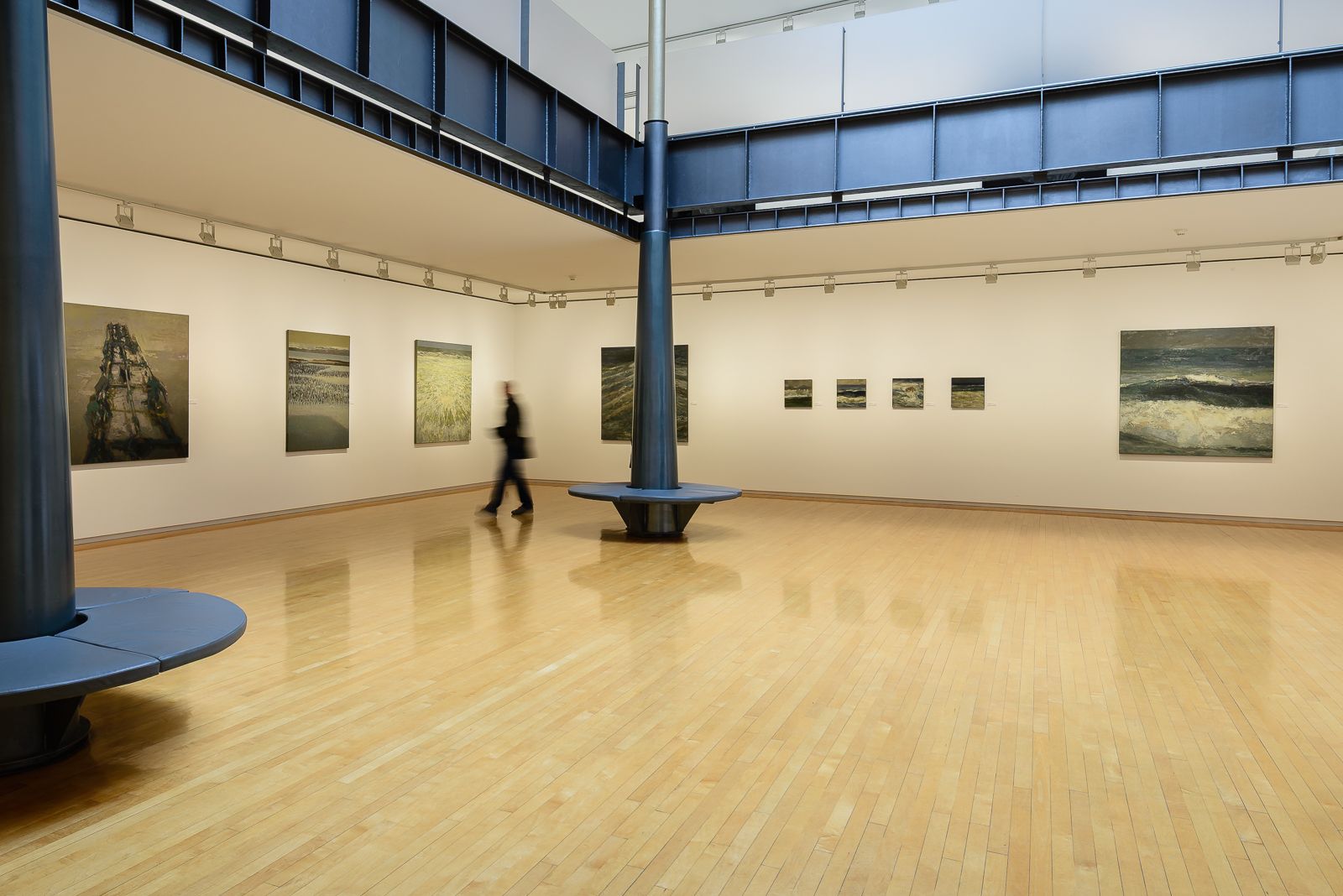 Galería de arte con suelos de madera, paredes blancas, pinturas de paisajes marinos y dos columnas negras con un banco circular, con una persona borrosa caminando al fondo.