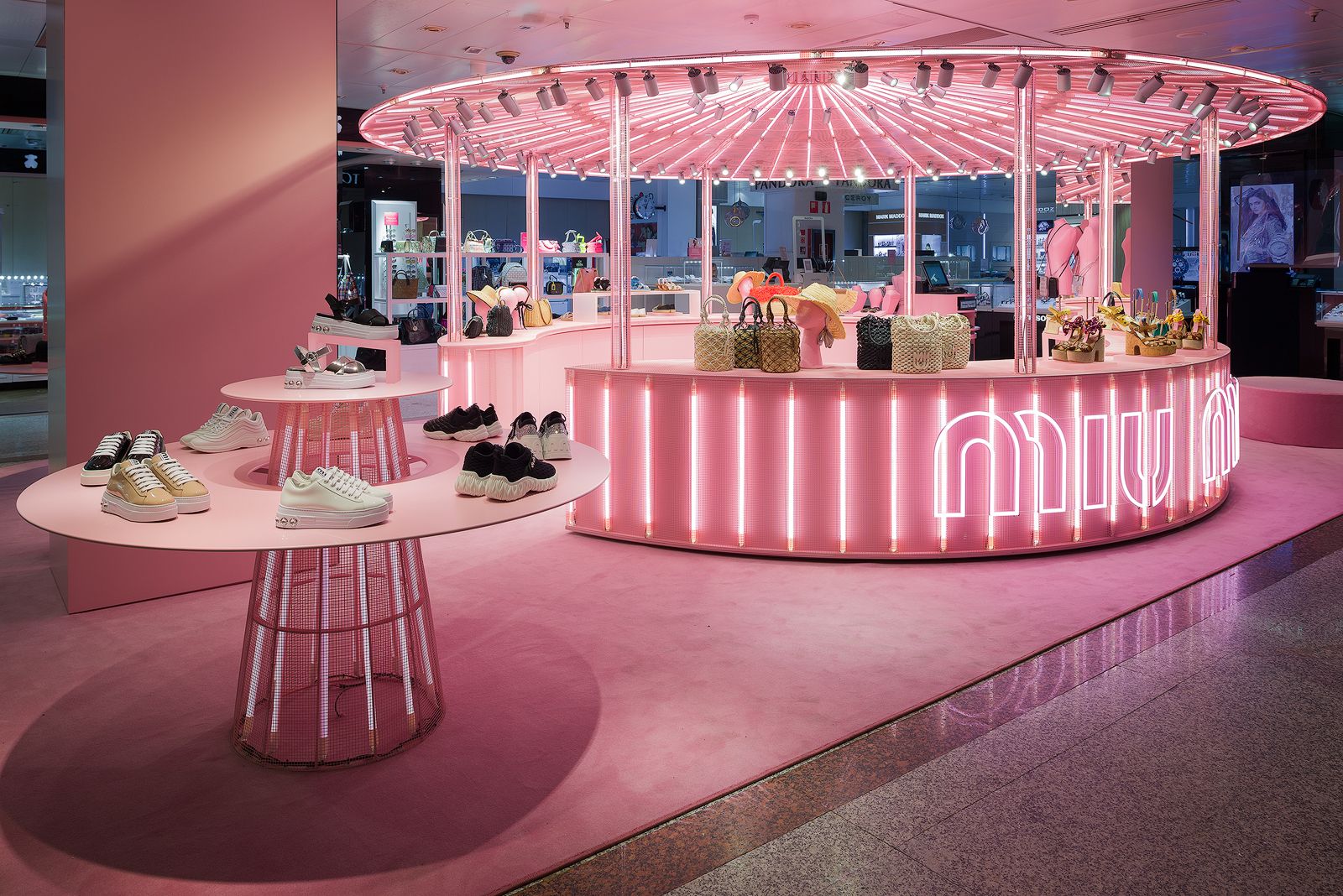 Exhibición de tienda pop-up de Miu Miu de temática rosa con mesas redondas iluminadas mostrando zapatillas, bolsos y accesorios.