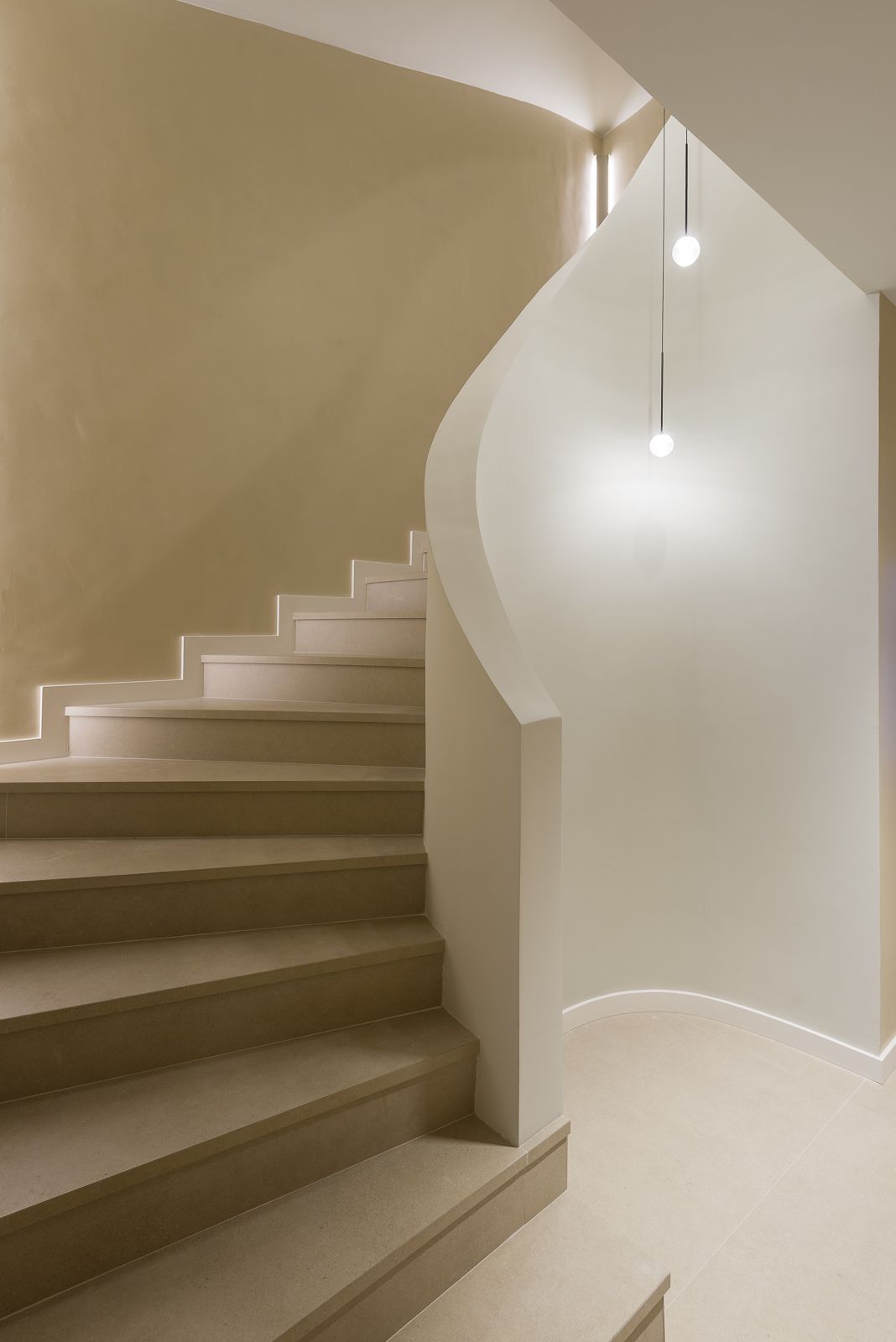 Modern minimalist interior featuring a beige staircase with stone steps and smooth plaster walls, bordered by a sculptural curved handrail and illuminated by two small glowing orb pendant lights hanging from a double-height ceiling.