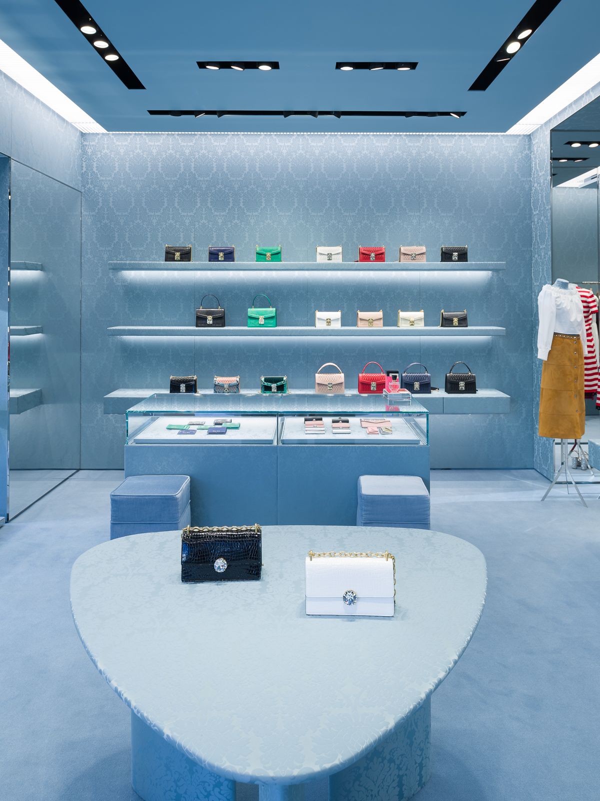 Interior de tienda Miu Miu con paredes estampadas azules, accesorios de iluminación negros y bolsos coloridos exhibidos en elegantes estantes y mostradores de vidrio.