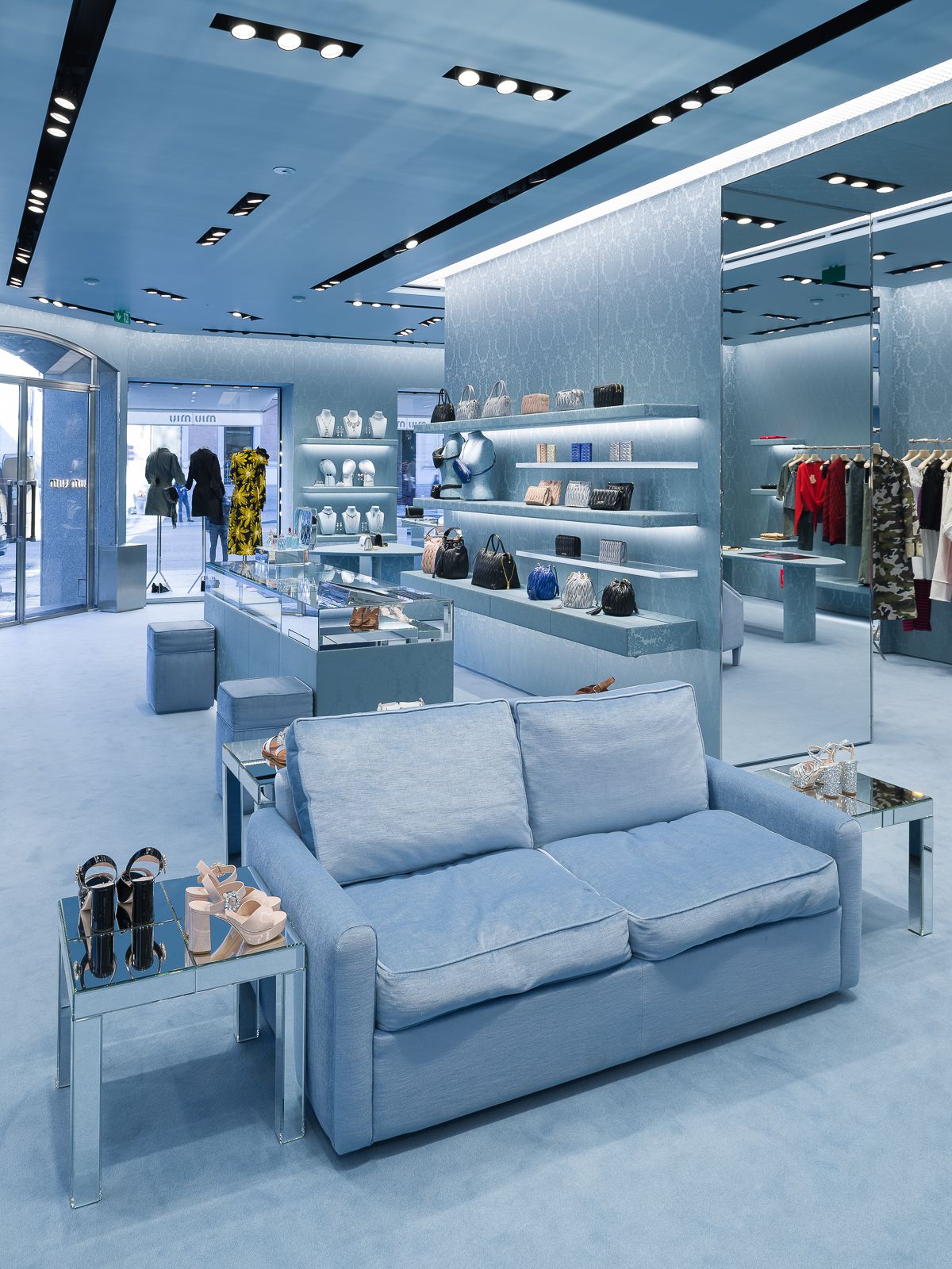 Interior de tienda Miu Miu con decoración azul, mostrando un área de asientos, estantes de exhibición con bolsos y accesorios, y un mostrador de exhibición de vidrio con zapatos.