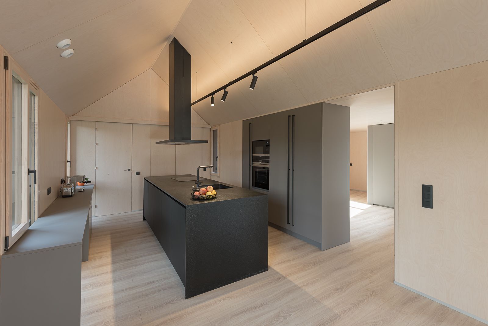 Vista interior de una cocina minimalista con una isla negra central, armarios elegantes y paredes y suelos de madera clara. El espacio está iluminado por iluminación de riel y luz natural de las ventanas.