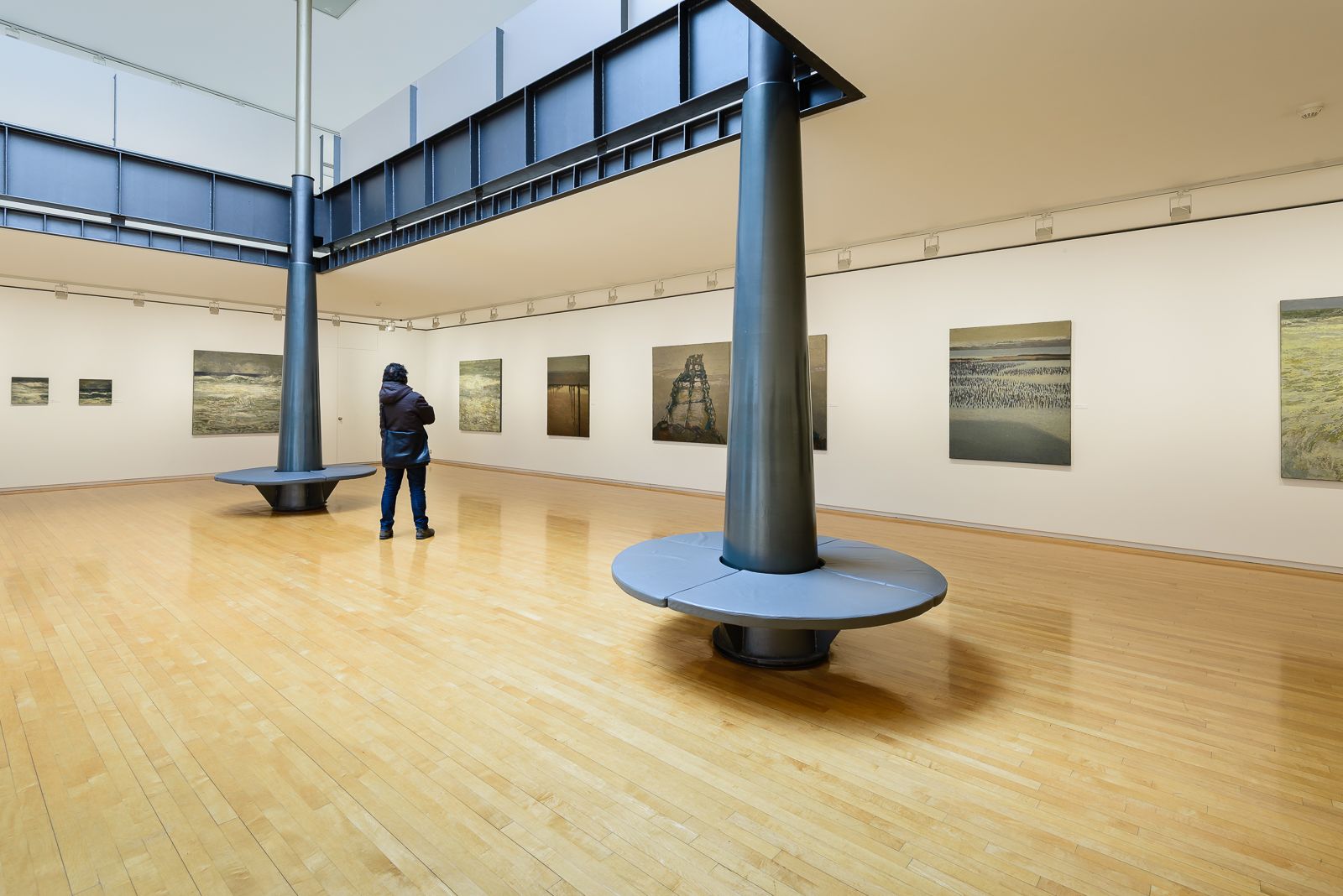 Galería de arte con suelos de madera, paredes blancas, pinturas de paisajes marinos y columnas negras, con una persona viendo las obras de arte.