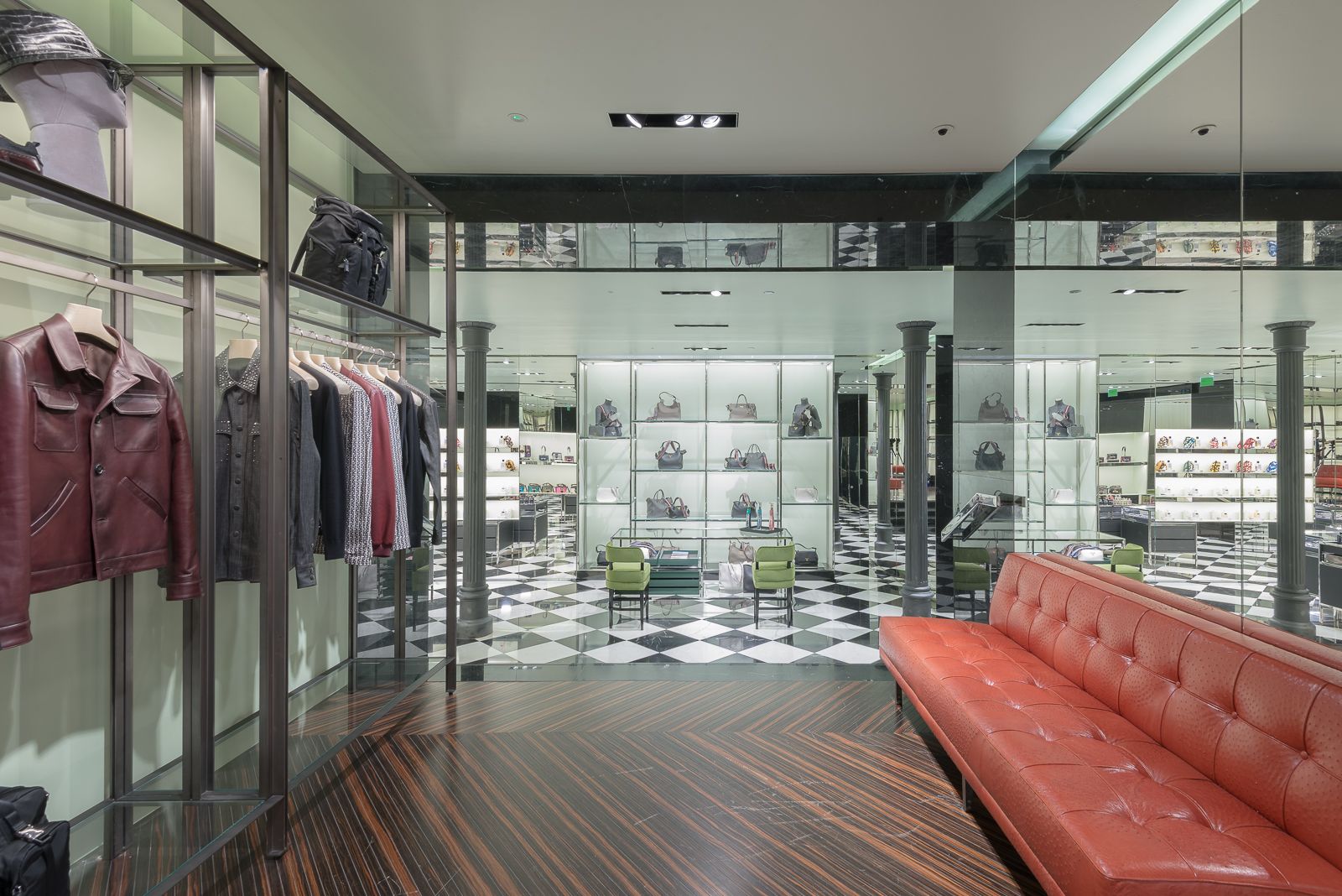 Interior de tienda Prada con percheros de ropa, chaquetas de cuero y suelo de tablero de ajedrez, con un sofá rojo y estantes de exhibición al fondo.