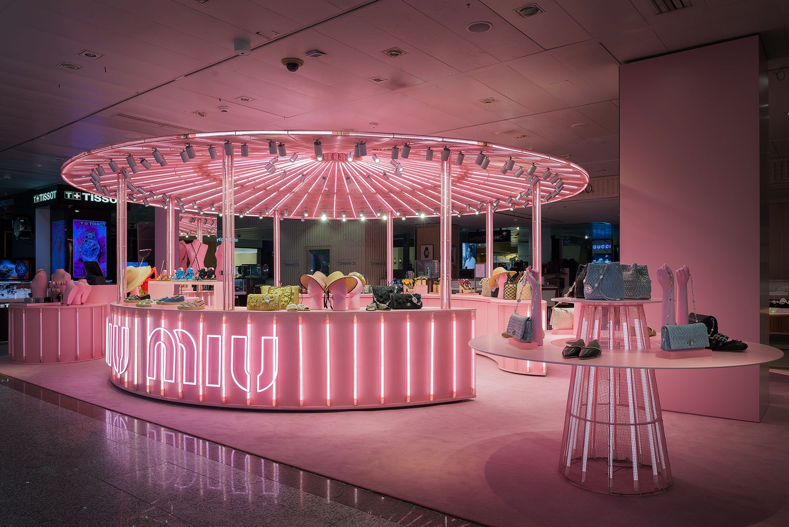 Tienda pop-up de Miu Miu de temática rosa con un área de exhibición circular, iluminada por vibrantes luces de neón rosas, mostrando varios bolsos y accesorios.
