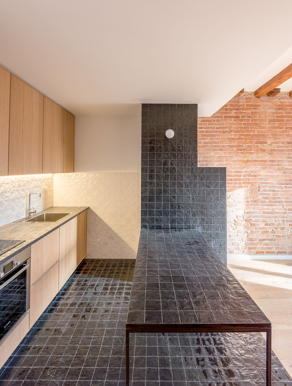 Cocina moderna con encimera y suelo de baldosas negras, armarios de madera y pared de ladrillo expuesto.