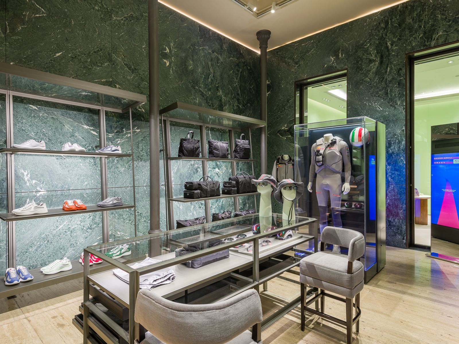 Interior de boutique de alta gama con paredes de mármol verde, elegantes estanterías metálicas que exhiben zapatillas y bolsos, y una vitrina de vidrio con un maniquí en un atuendo de rendimiento con un casco de carreras. Una mesa de vidrio central contiene accesorios, con asientos acolchados cerca.