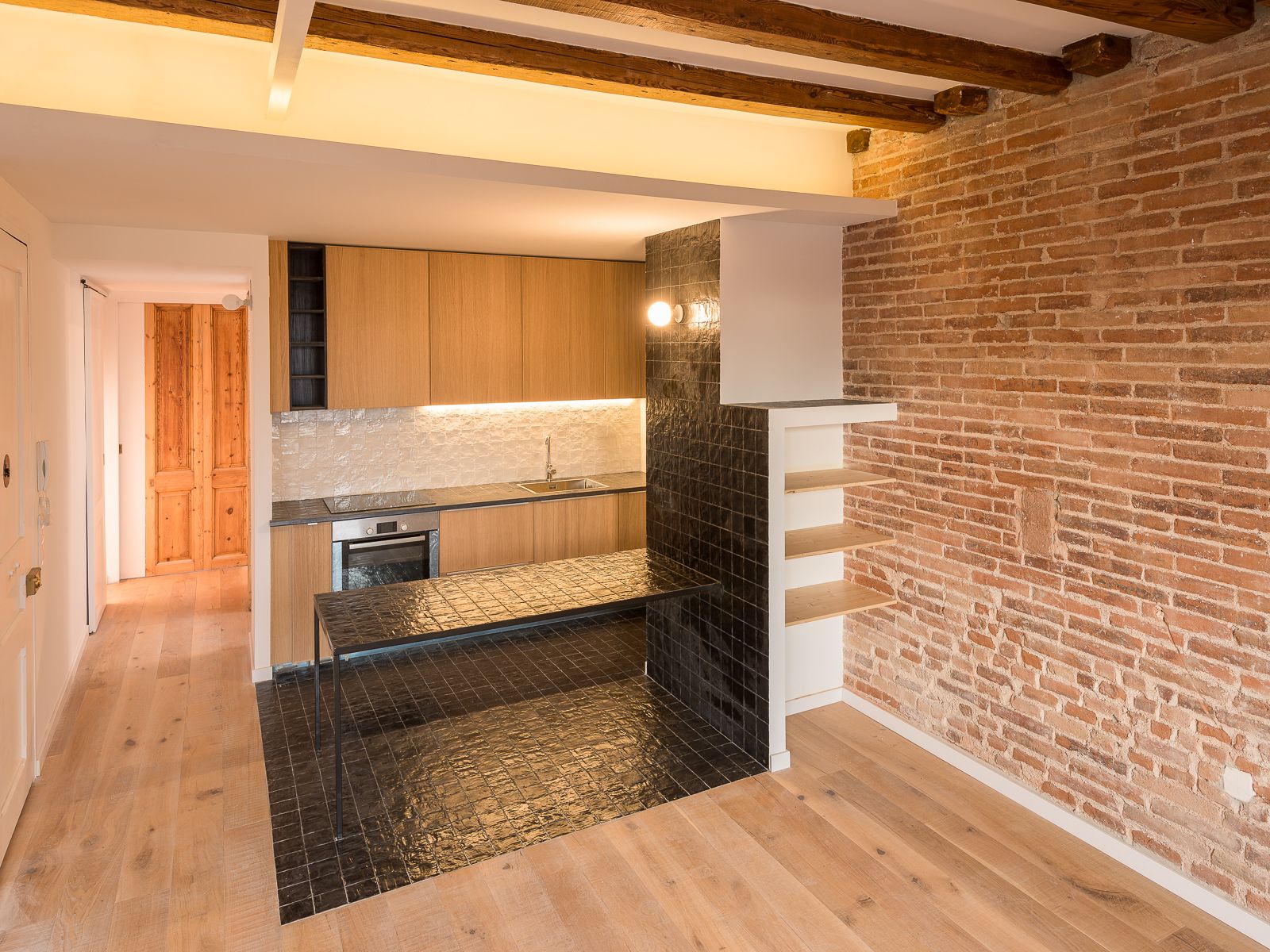 Cocina abierta moderna con paredes de ladrillo expuesto, suelos de madera clara y encimera de baldosas negras con mesa de trabajo de baldosas negras bajo techo de vigas de madera.