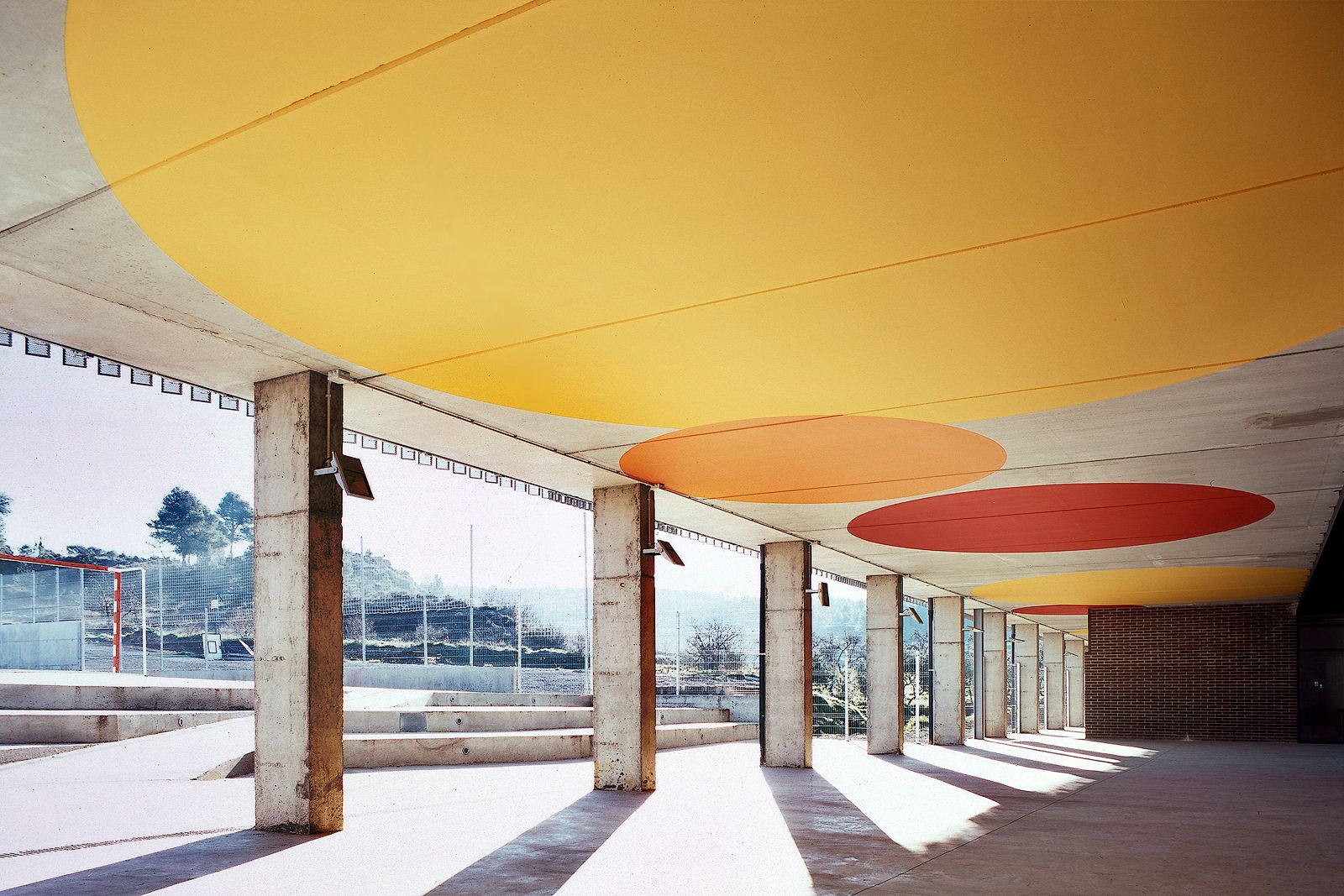 A low-angle architectural photograph of a covered outdoor school pavilion. The perspective follows a row of exposed concrete pillars casting sharp shadows onto the ground. The underside of the concrete roof features a striking geometric design with large, vibrant circles painted in yellow, orange, and red. In the background, concrete bleachers lead up to an outdoor sports court with a wire fence under a bright sky.