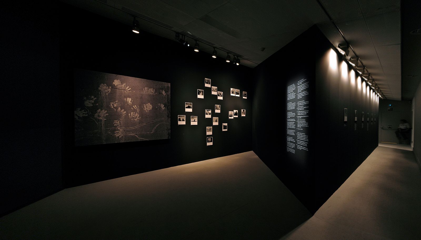 * Fotografía panorámica de una sala de exposiciones de arte con iluminación tenue y paredes de color negro mate, que muestra una gran obra con huellas de manos, un conjunto de pequeñas fotografías y un pasillo contiguo iluminado.