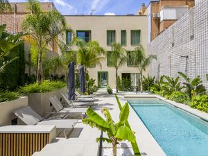 Pati exterior lluminós amb una piscina de natació d'aigua transparent, un pati de pedra beix amb gandules i para-sols, i una exuberant vegetació tropical que inclou palmeres i plataners, amb l'exterior d'un edifici modern de tres plantes amb detalls verds de fons.