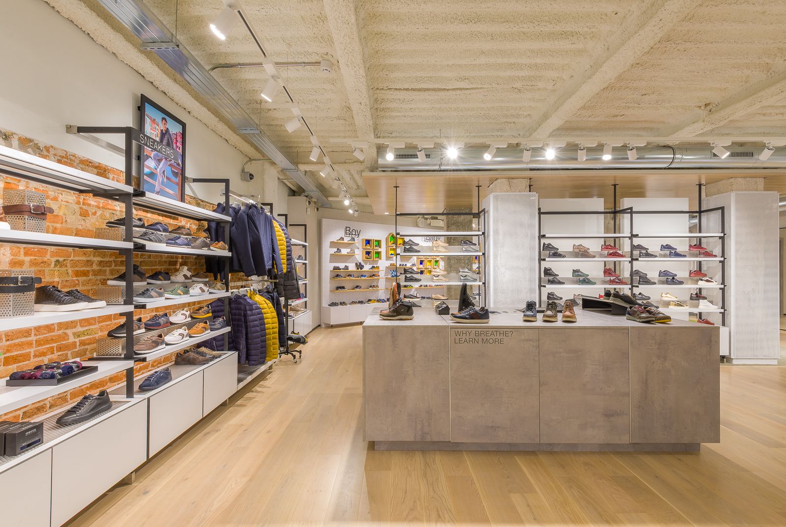 Vista interior de la tienda Geox con estantes de zapatos y chaquetas contra una pared de ladrillo expuesto, una mesa de exhibición con varios zapatos y estantes de zapatos adicionales al fondo.