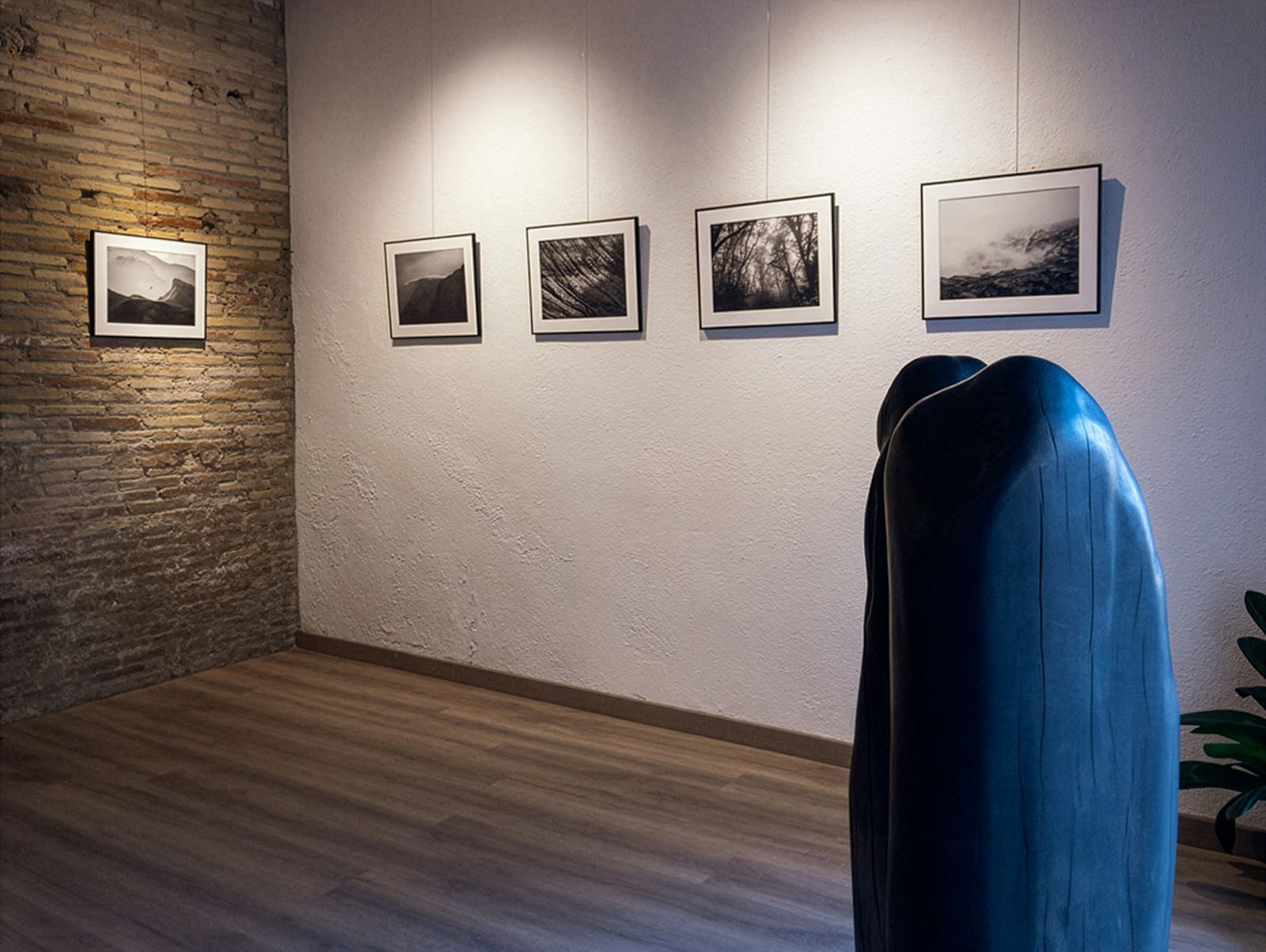 Exposición de fotografía con fotos de paisajes en blanco y negro en paredes blancas y de ladrillo, con una escultura oscura en primer plano.