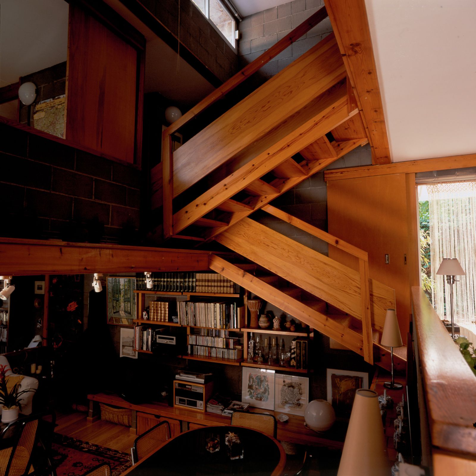 Vista interior con una escalera de madera, un área de comedor con sillas, estanterías llenas de libros y objetos decorativos, y un ambiente acogedor creado por la combinación de elementos de madera y paredes de bloques de hormigón.
