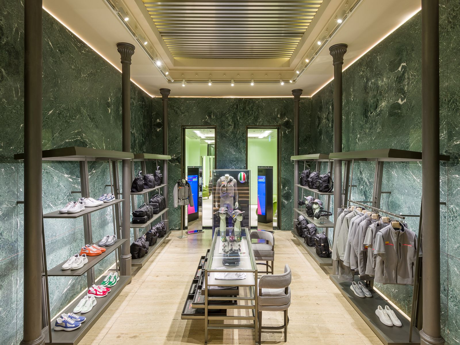 Lujoso interior de boutique con paredes de mármol verde, elegante iluminación y estanterías metálicas negras que exhiben zapatillas, bolsos y prendas grises. Una mesa de vidrio central muestra accesorios, mientras que una puerta al fondo conduce a otra sección.