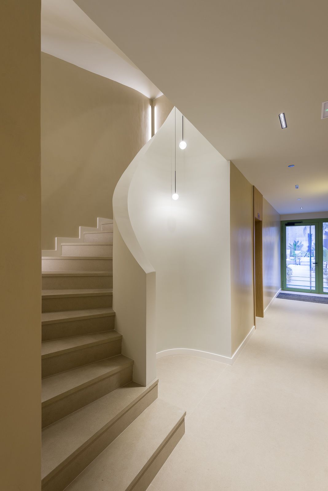 Wide-angle interior view of a modern, minimalist building entryway featuring a beige stone staircase with a sweeping, sculptural curved wall, soft ambient lighting from two delicate globe pendant lights, and a long hallway leading past an elevator toward a glass exit door with green trim.