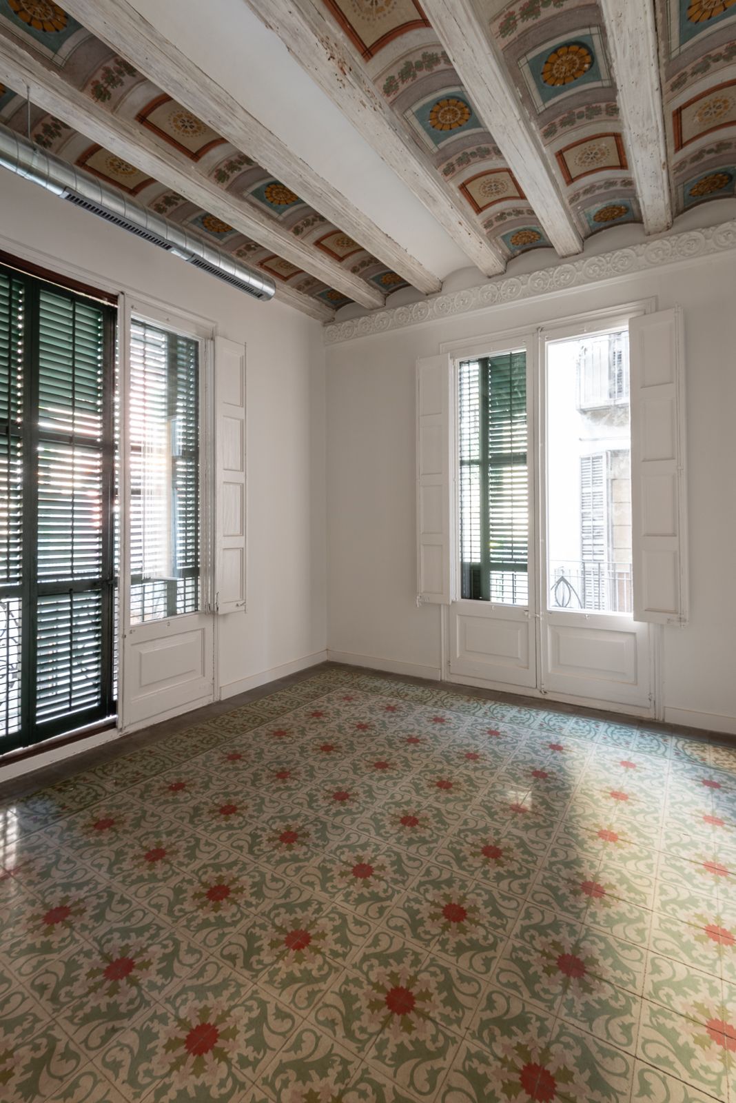 Habitación con baldosas de suelo estampadas, techo con vigas ornamentadas y grandes ventanas con contraventanas.