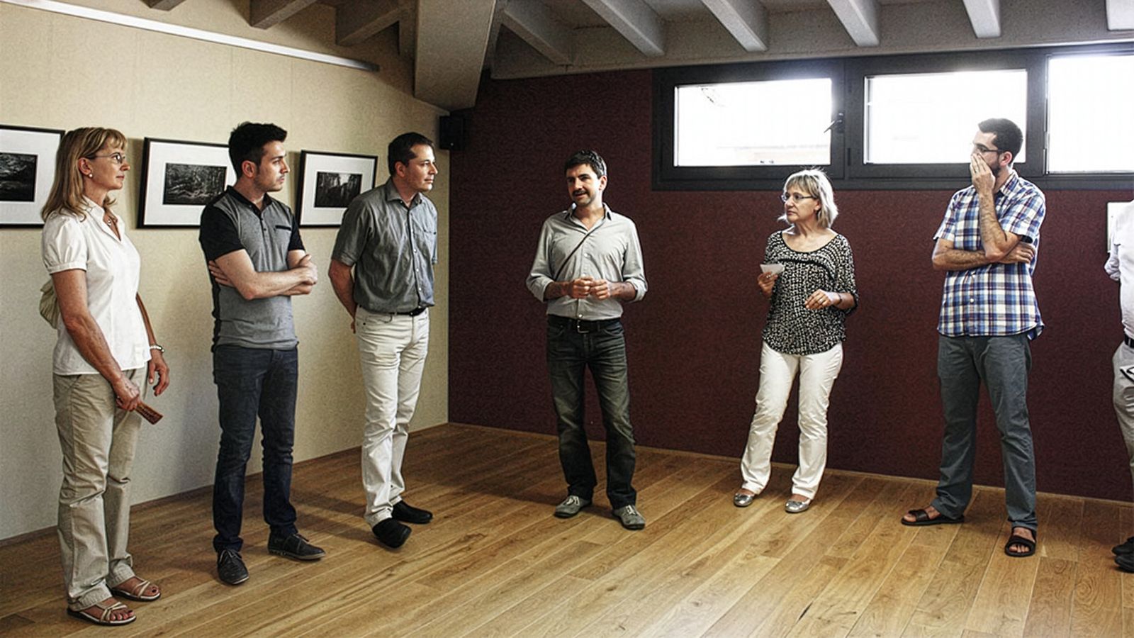 Grupo de personas de pie en una galería de arte, escuchando a un hombre hablar.
