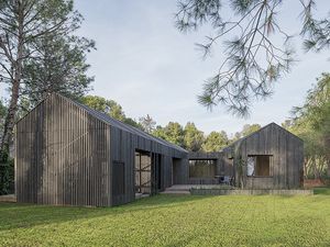 Vista exterior de una casa moderna con dos cuerpos, techo inclinado, revestimiento de madera oscura, grandes ventanas y un amplio césped rodeado de pinos.