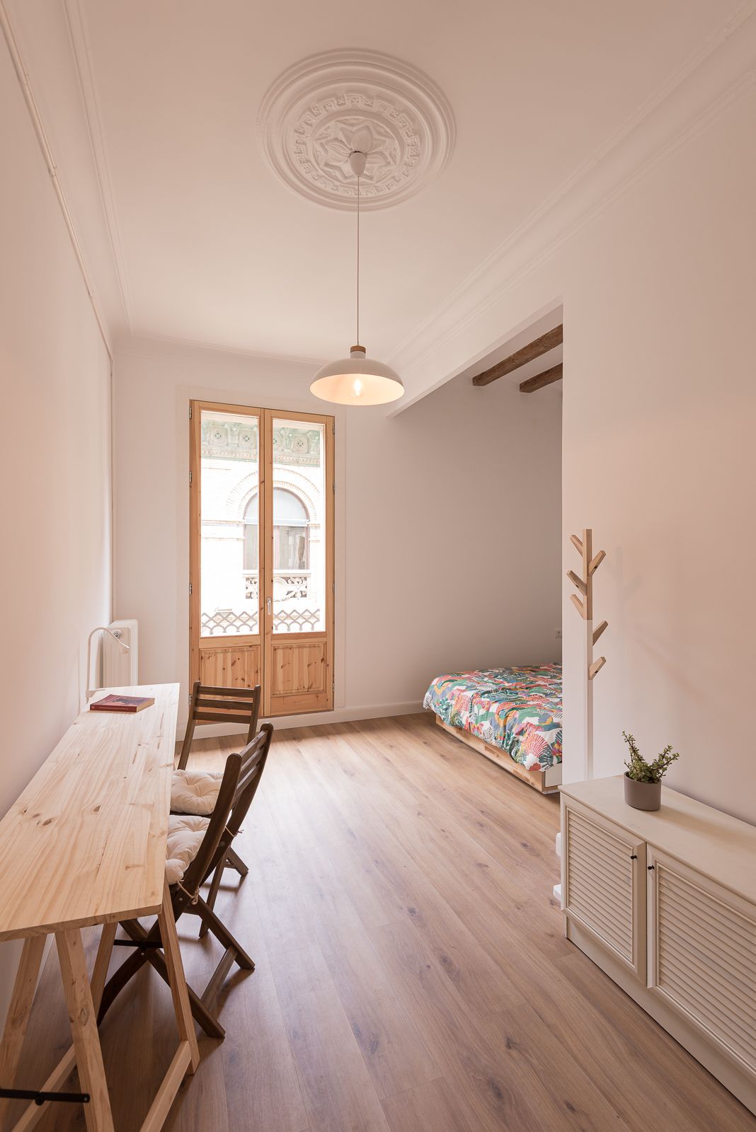 Bright room with wooden floors, a studio desk, and a view into a colorful bed and a balcony.
