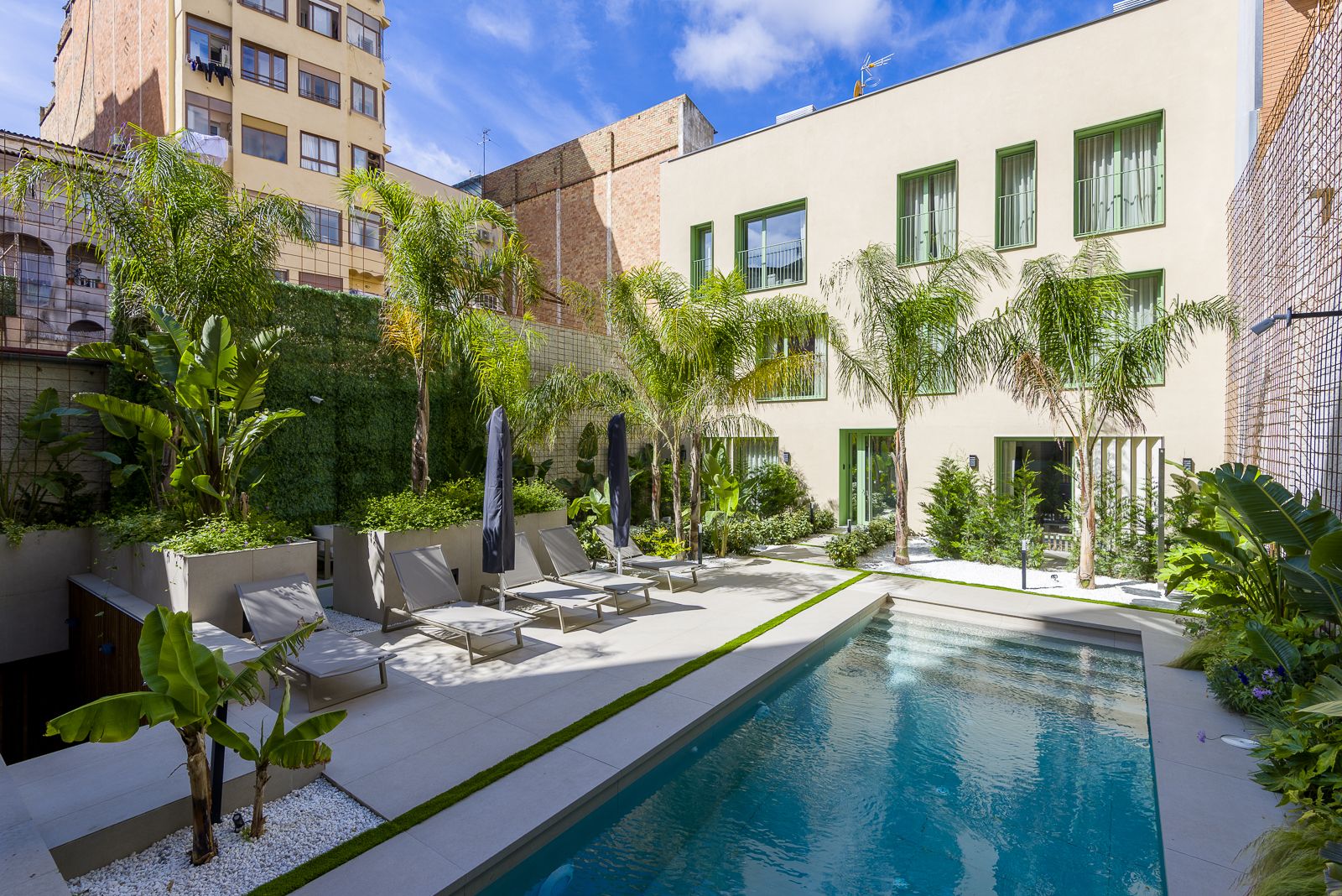 Sunny urban courtyard garden featuring a rectangular swimming pool surrounded by beige pavers, lounge chairs, palm trees, and lush tropical foliage, set against the rear facade of a modern light beige three-story building with green window trim.