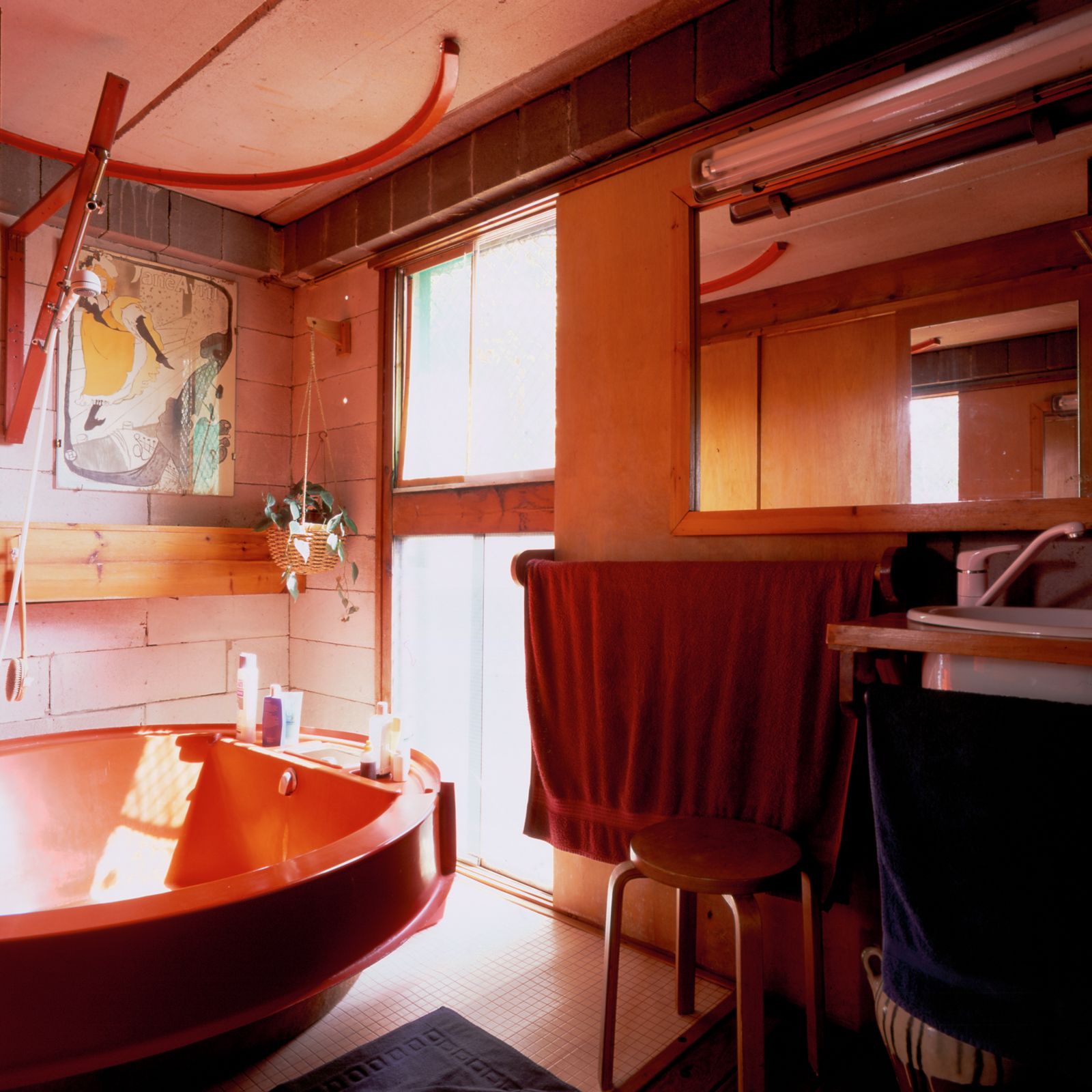Baño con bañera roja, elementos de madera, ventana de suelo a techo y una variedad de toallas colgando debajo de un espejo y un lavabo.