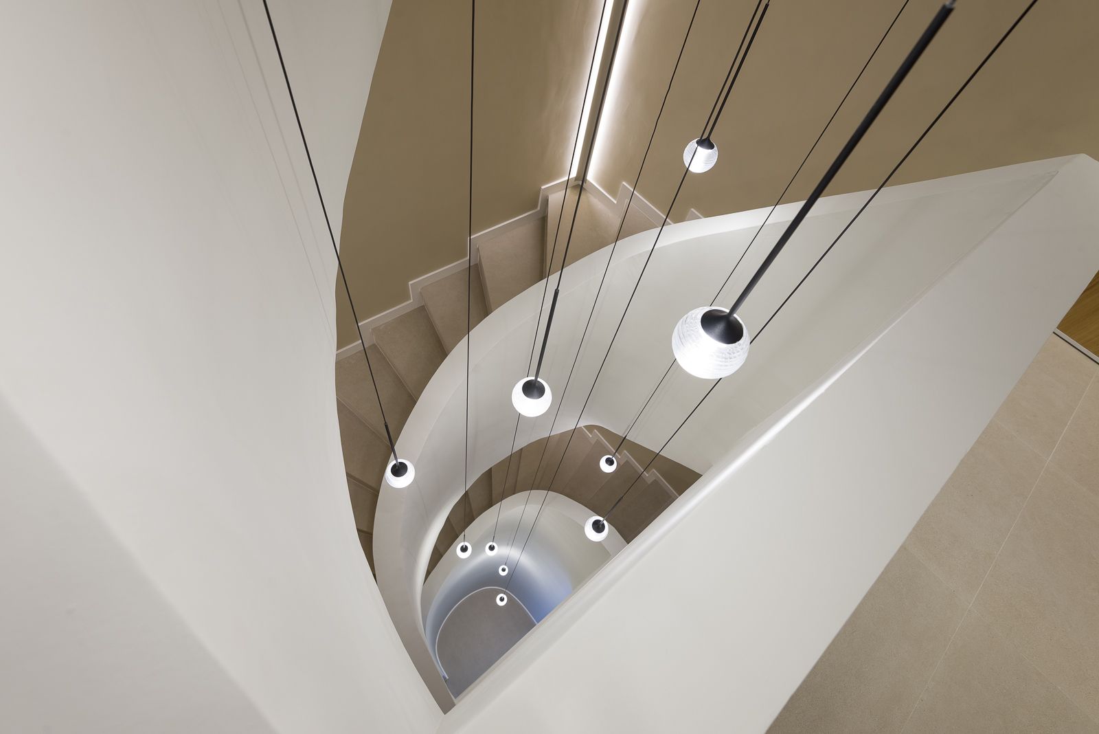High-angle downward view into the central void of a smooth beige spiral staircase, showing sweeping curved balustrades, integrated recessed lighting along the steps, and a cascade of globe pendant lights suspended on thin black cords.
