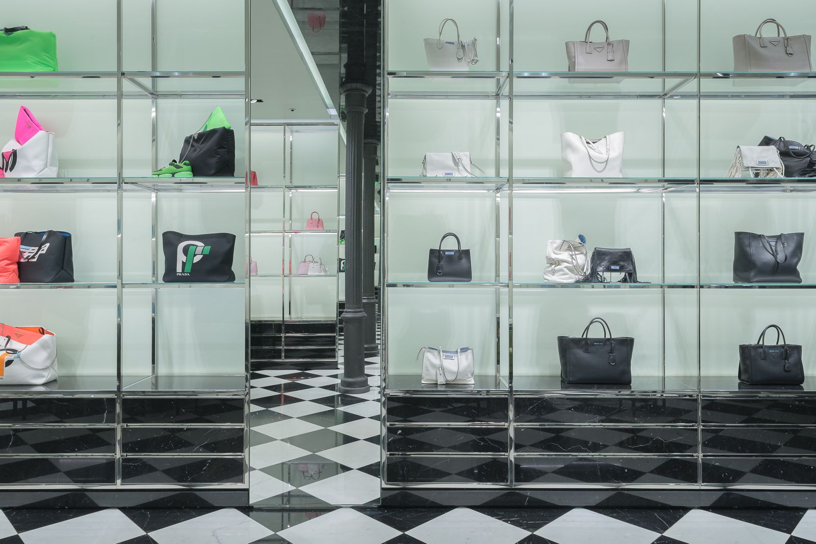 Estantes de bolsos de alta gama en varios colores exhibidos contra un fondo verde claro en una tienda Prada, con dos espejos enfrentados y una columna de hierro fundido negro atrapada en el efecto de espejo infinito.