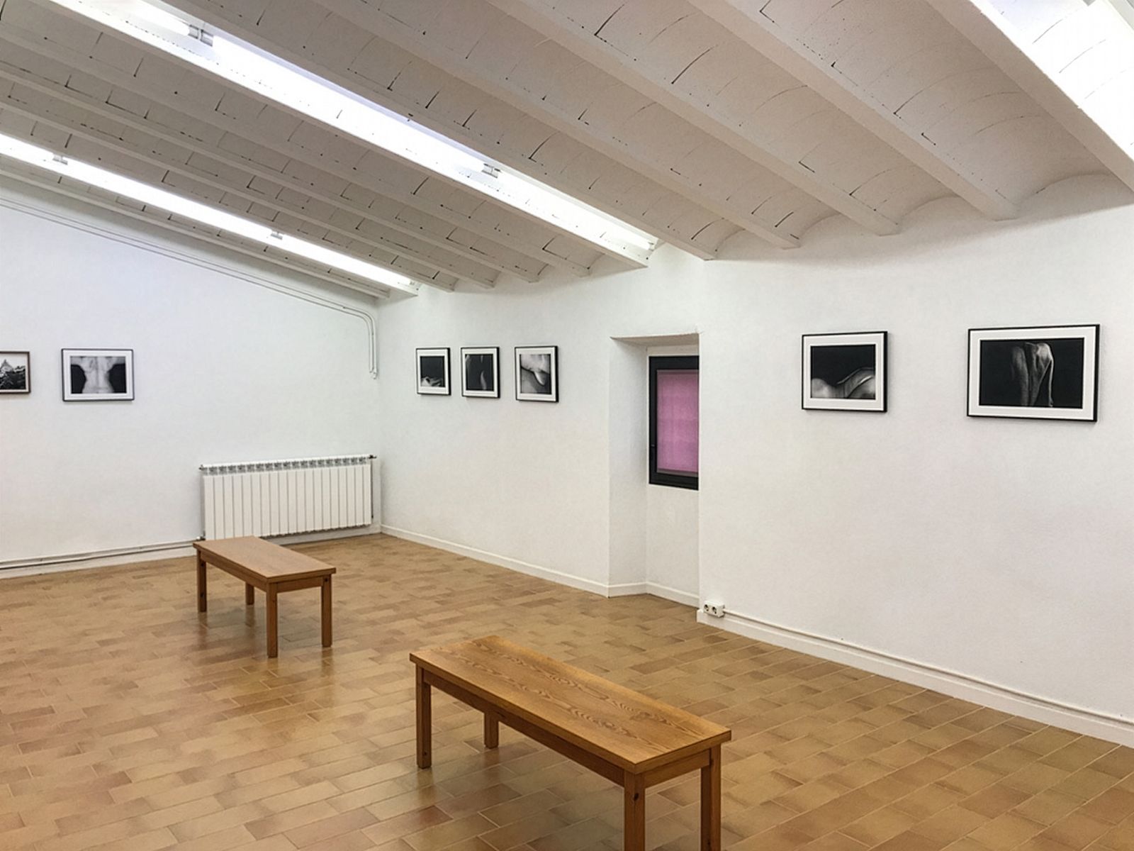Una sala de galeria minimalista amb fotografies en blanc i negre en parets blanques, dos bancs de fusta al centre i una petita finestra en una paret.