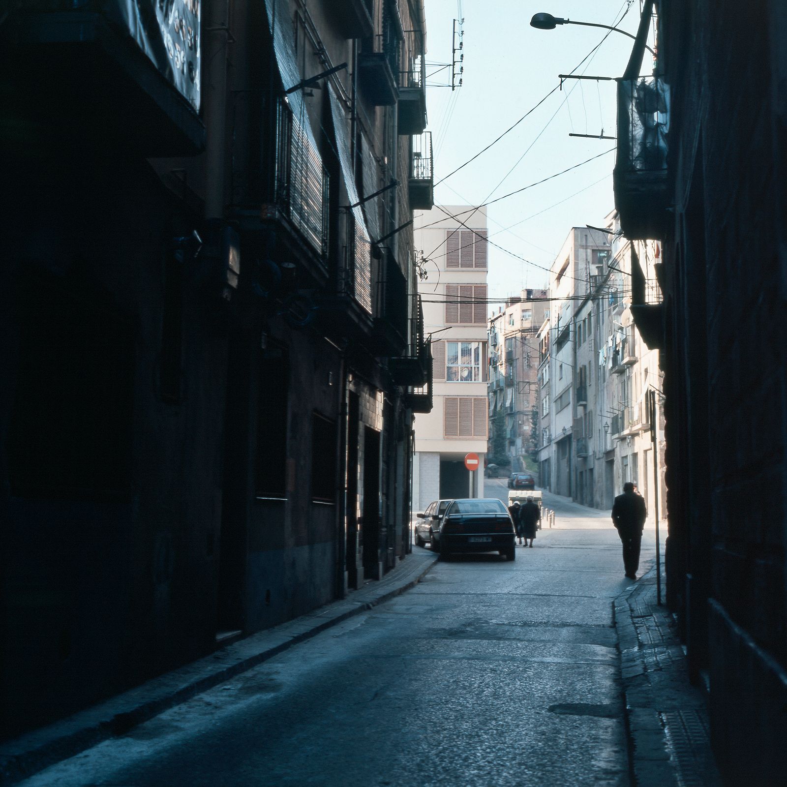 Un callejón estrecho con edificios en sombra a ambos lados, que conduce a una plaza más luminosa y un nuevo edificio residencial al fondo con algunos peatones y coches aparcados.
