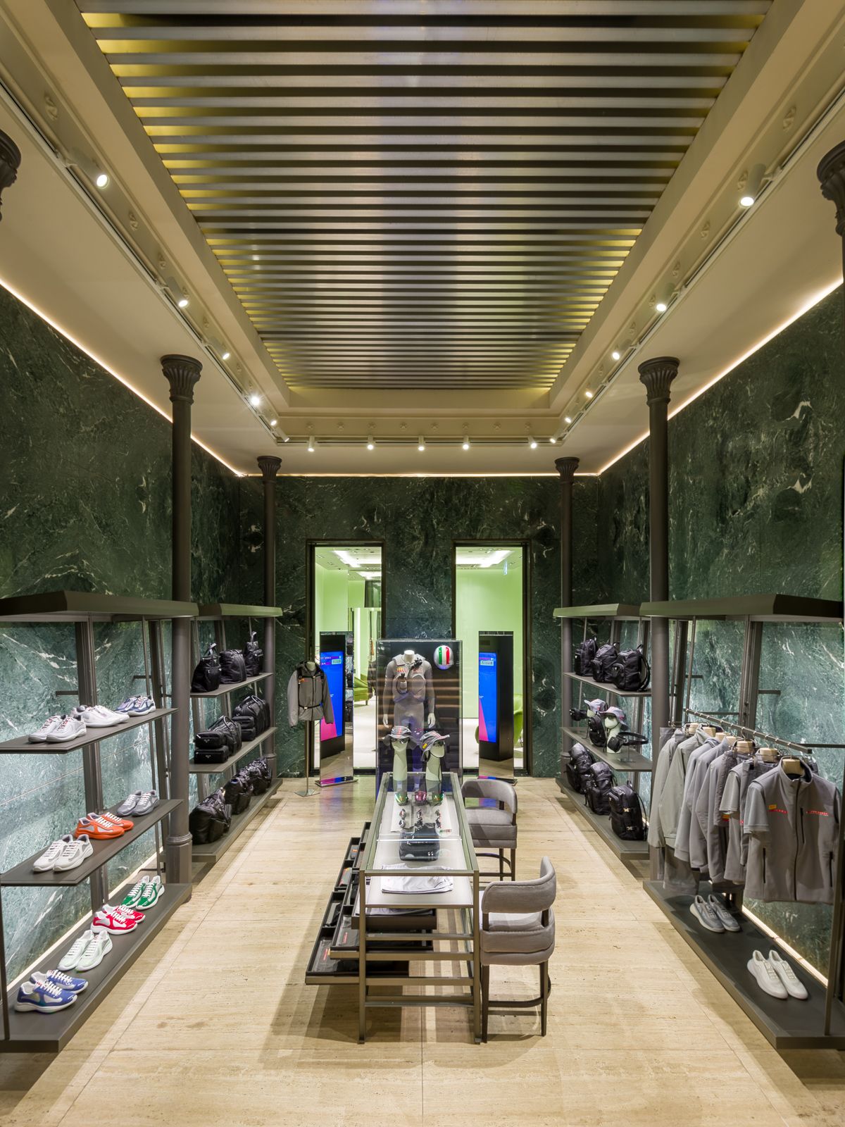 Lujoso interior de boutique con paredes de mármol verde, elegante iluminación y estanterías metálicas negras que exhiben zapatillas, bolsos y prendas grises. Una mesa de vidrio central muestra accesorios, mientras que una puerta al fondo conduce a otra sección.