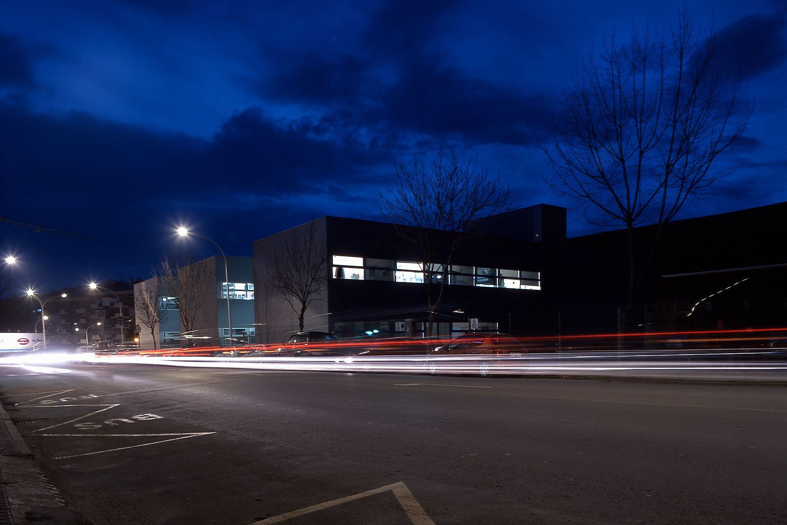 Nighttime street scene with a modern building in the background, car light trails, and streetlights illuminating the area.