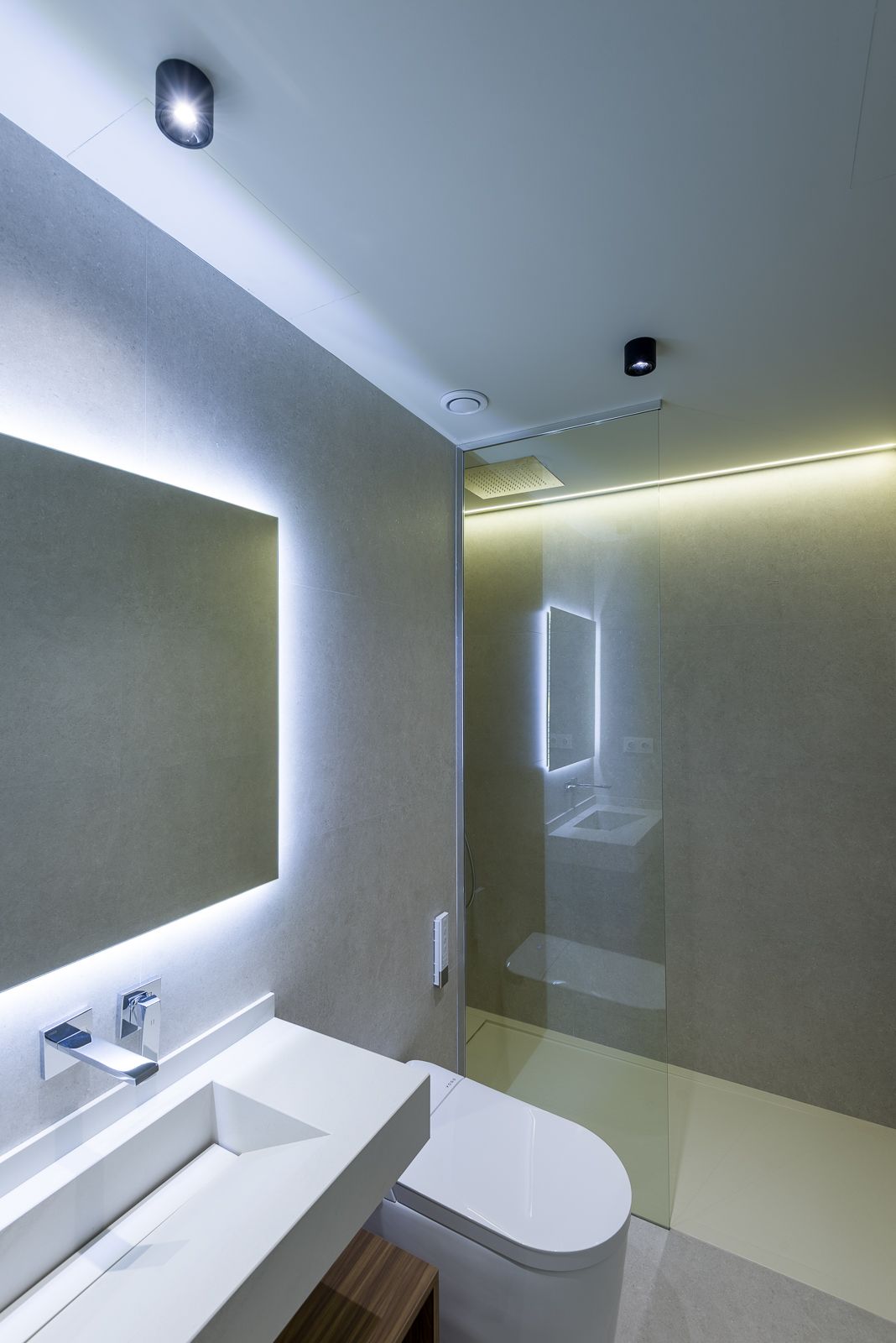 Modern minimalist bathroom with smooth grey concrete-look walls, a white floating vanity with wall-mounted chrome fixture, a back-lit rectangular mirror, and a glass-enclosed walk-in shower with indirect LED lighting.