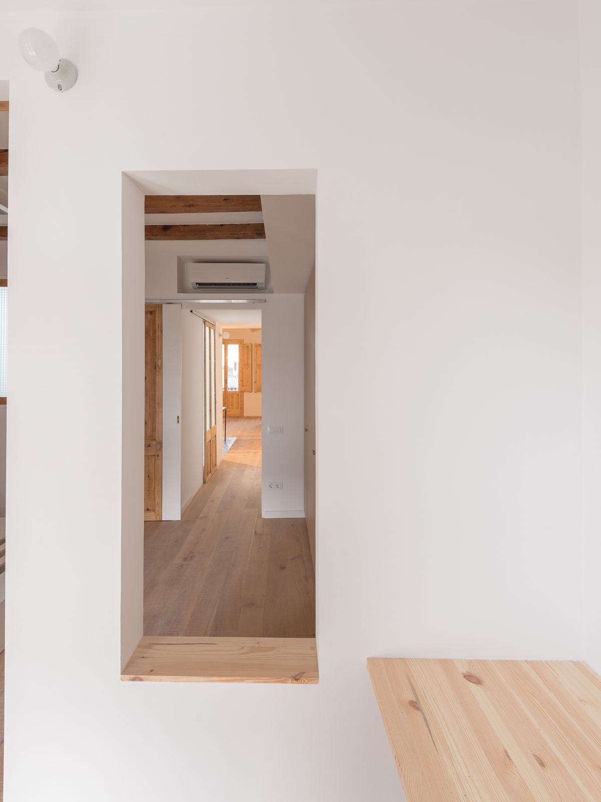 Una vista desde la habitación de estudio a través de una ventana abierta hacia una habitación con suelos de madera y paredes blancas, al fondo un pasillo que conduce al área de estar principal.
