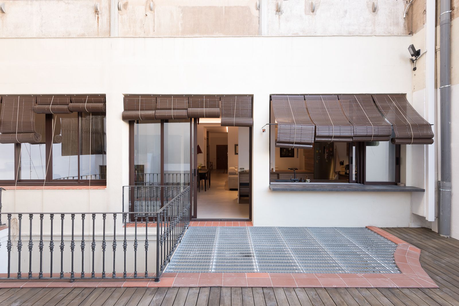 Outdoor patio with metal railing, wooden deck, and large windows with bamboo shades, leading into a modern interior.