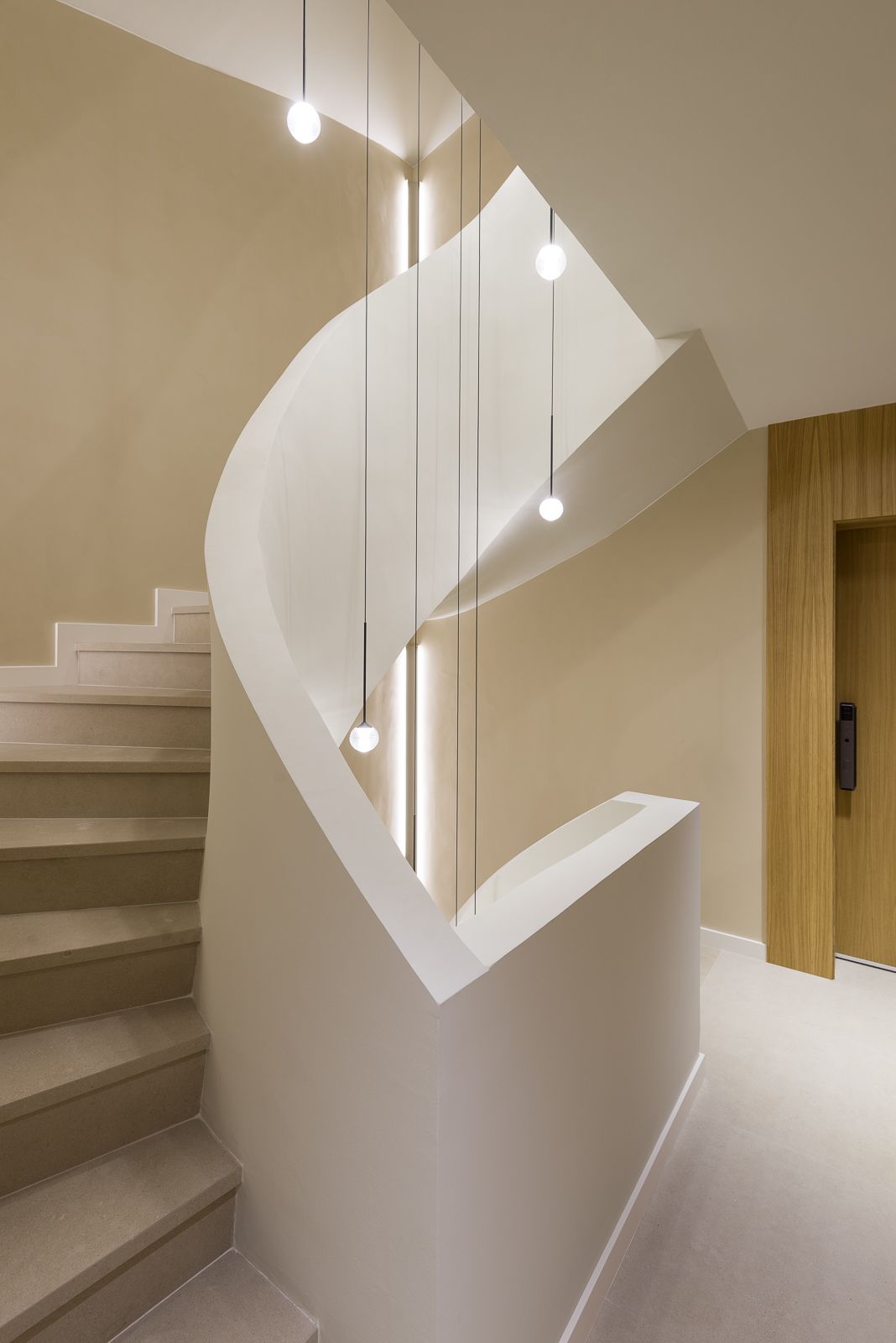 Architectural view looking up a modern, multi-story spiral staircase with smooth beige plaster walls, a sweeping curved balustrade, and slender black wires suspending glowing orb pendant lights through the central stairwell alongside an elevator entrance framed in warm light wood.
