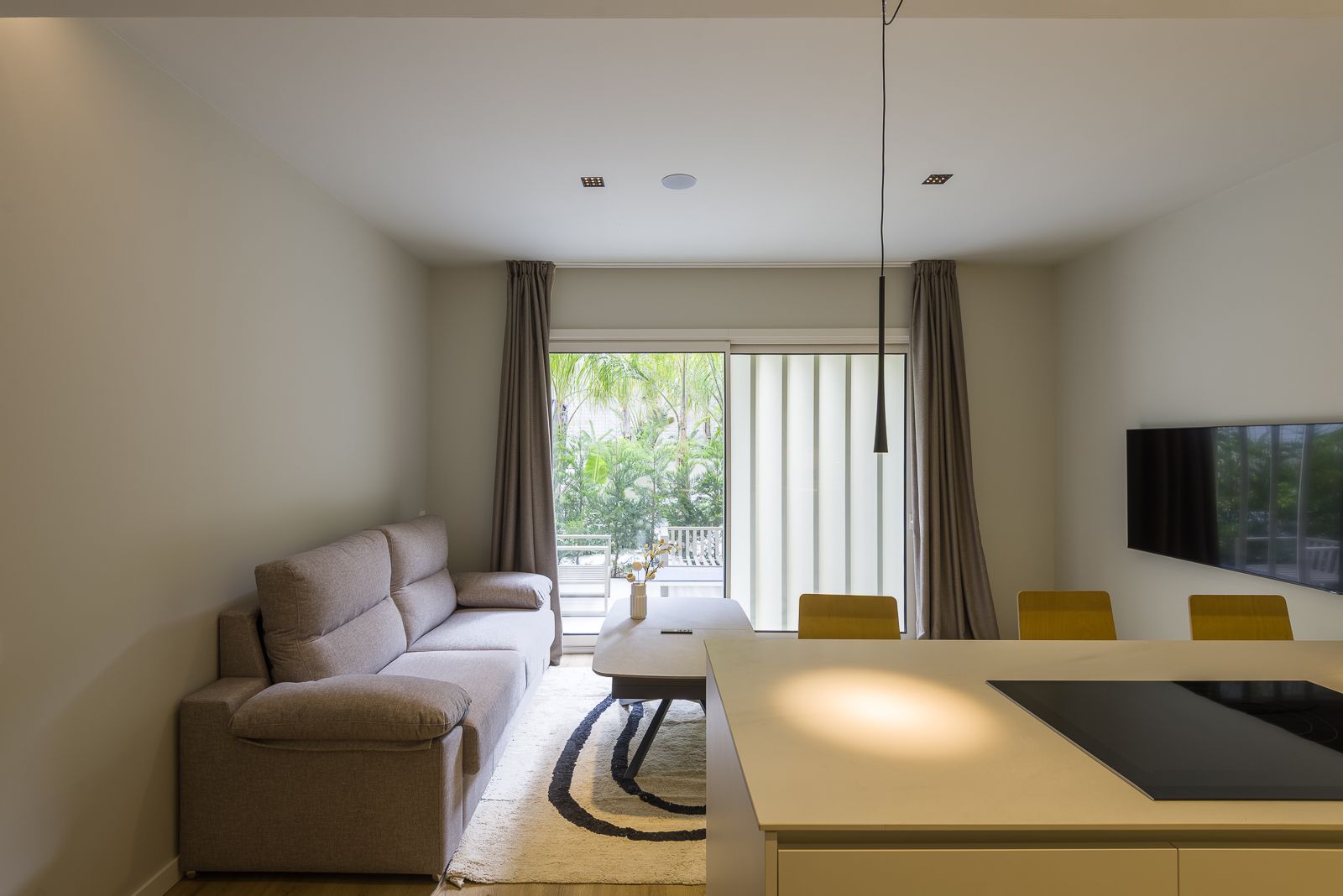 Modern open-plan living room viewed from a white kitchen counter, featuring a plush grey sofa, a patterned area rug, a wall-mounted TV, and large glass sliding doors framing an outdoor view of lush green trees.